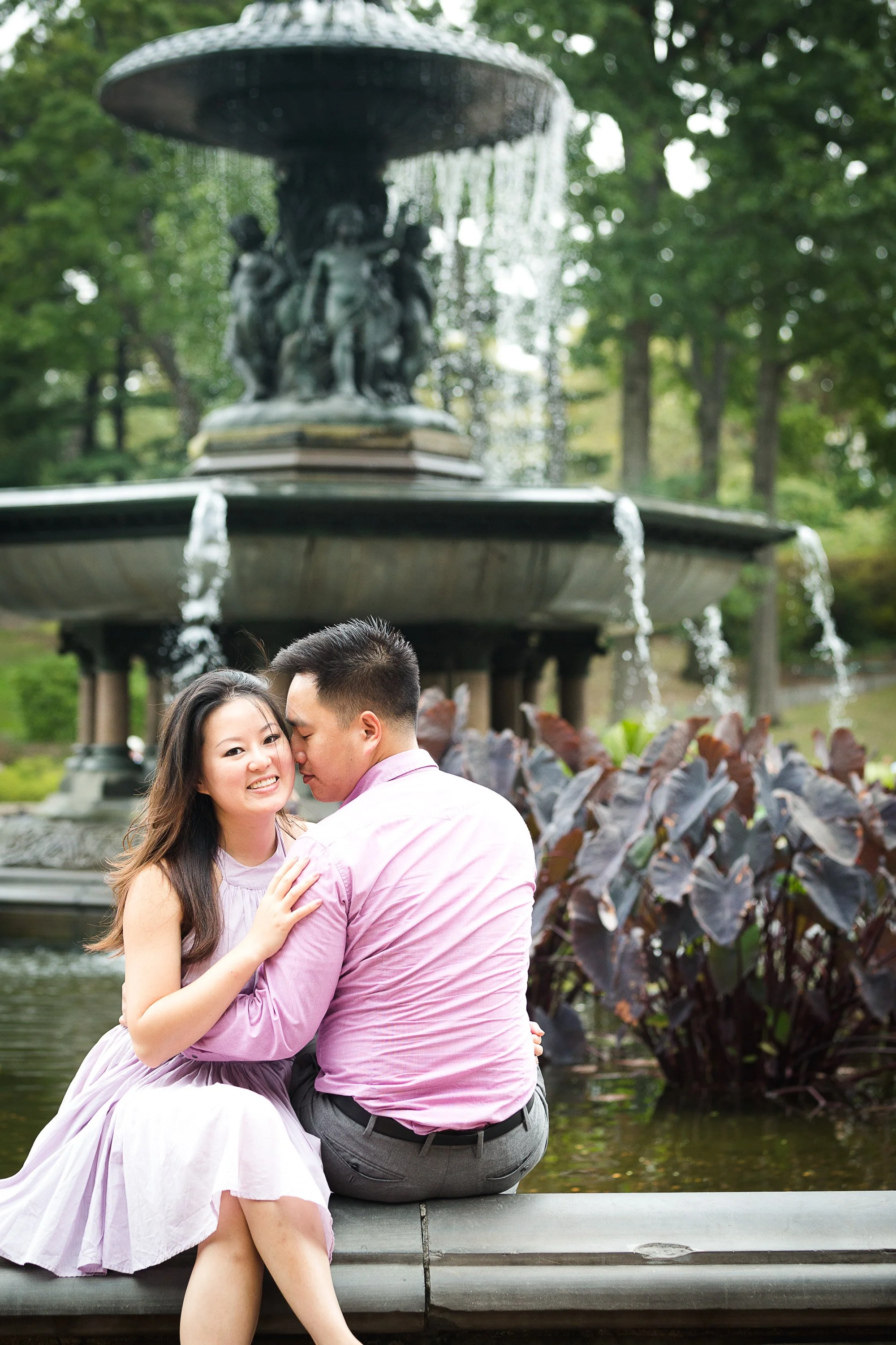 A couple sitting on a ledge in front of a fountain in a park, embracing and smiling, with trees in the background.