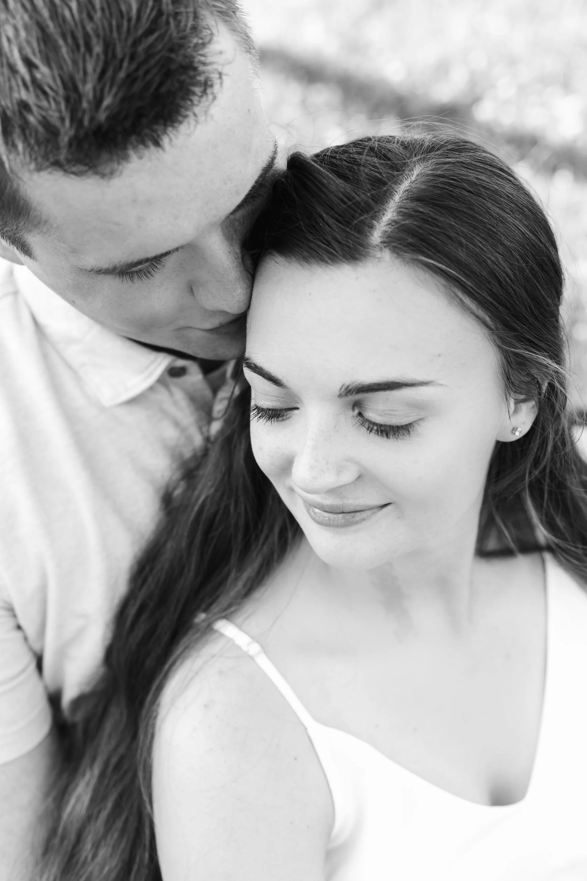 A black and white photo of a man and woman close together, with the man kissing the woman's forehead. The woman has closed eyes and a gentle smile, with long hair and wearing a sleeveless top.