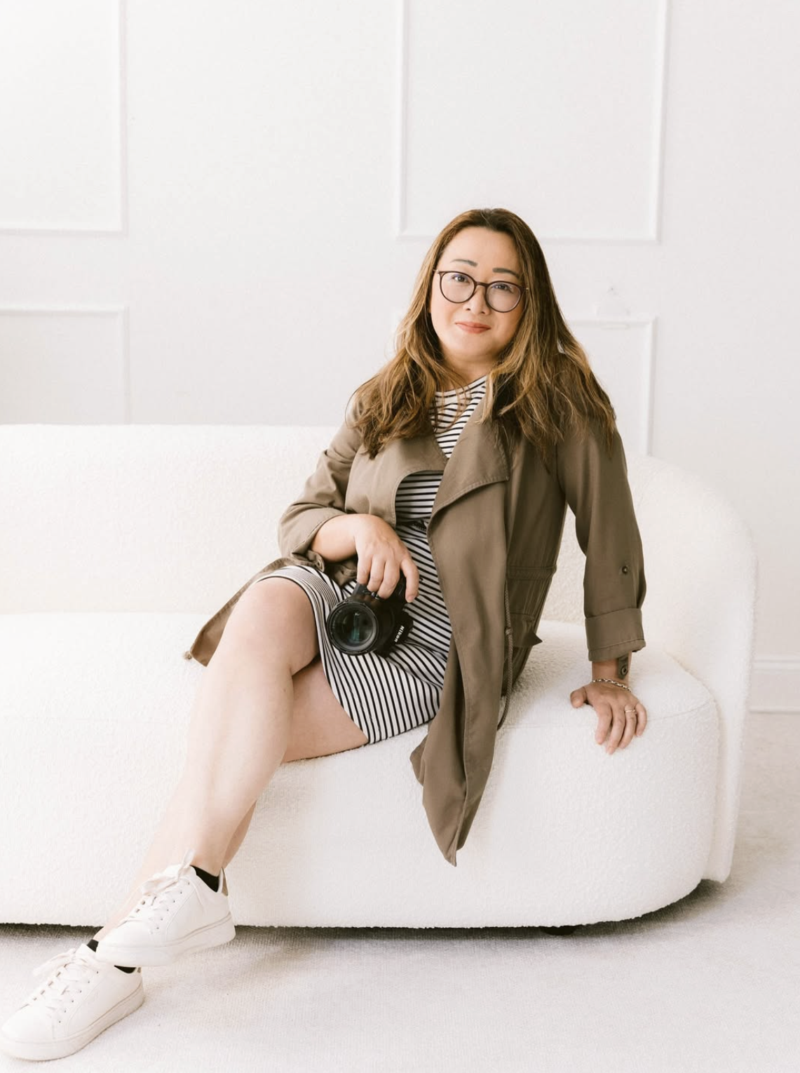 Woman sitting on a white couch, wearing glasses, a striped dress, white sneakers, and holding a camera, with a beige jacket draped over her shoulders.