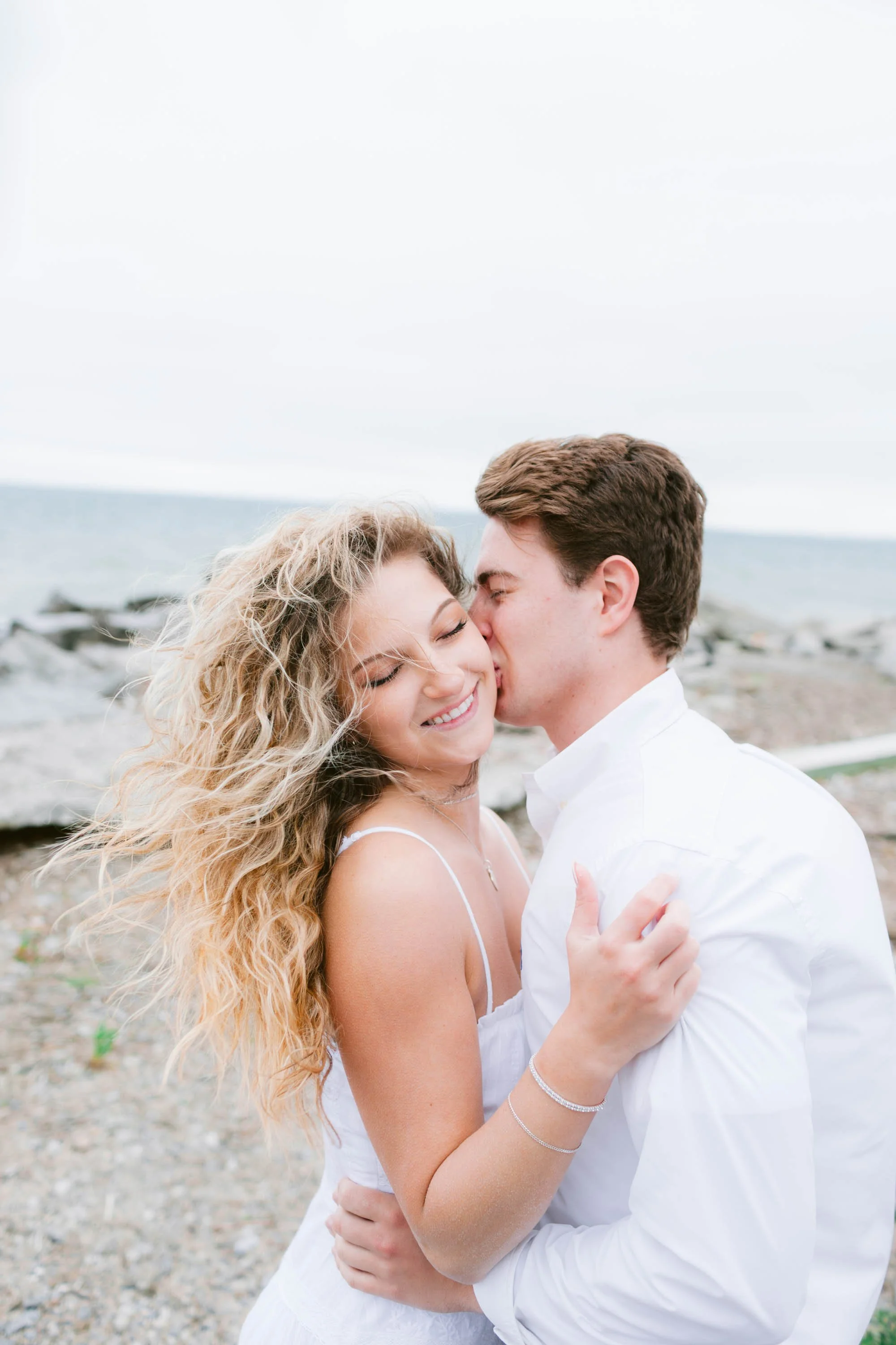 A couple hugging and sharing a kiss on a rocky beach with the ocean in the background.