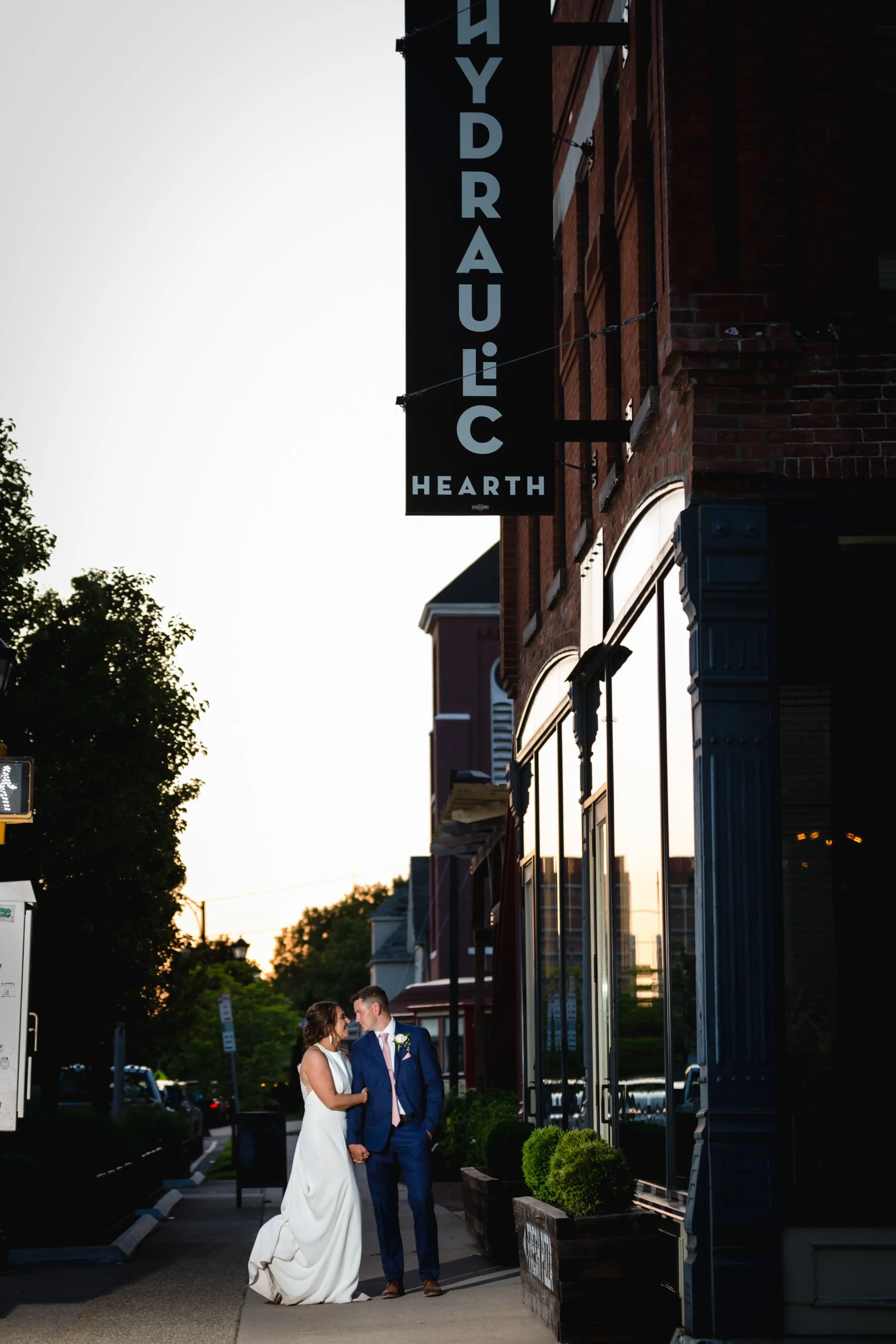 A newlywed couple stands outside a building during sunset, holding hands and gazing at each other, with a sign that reads 'Hydraulic Hearth' partially visible above them.