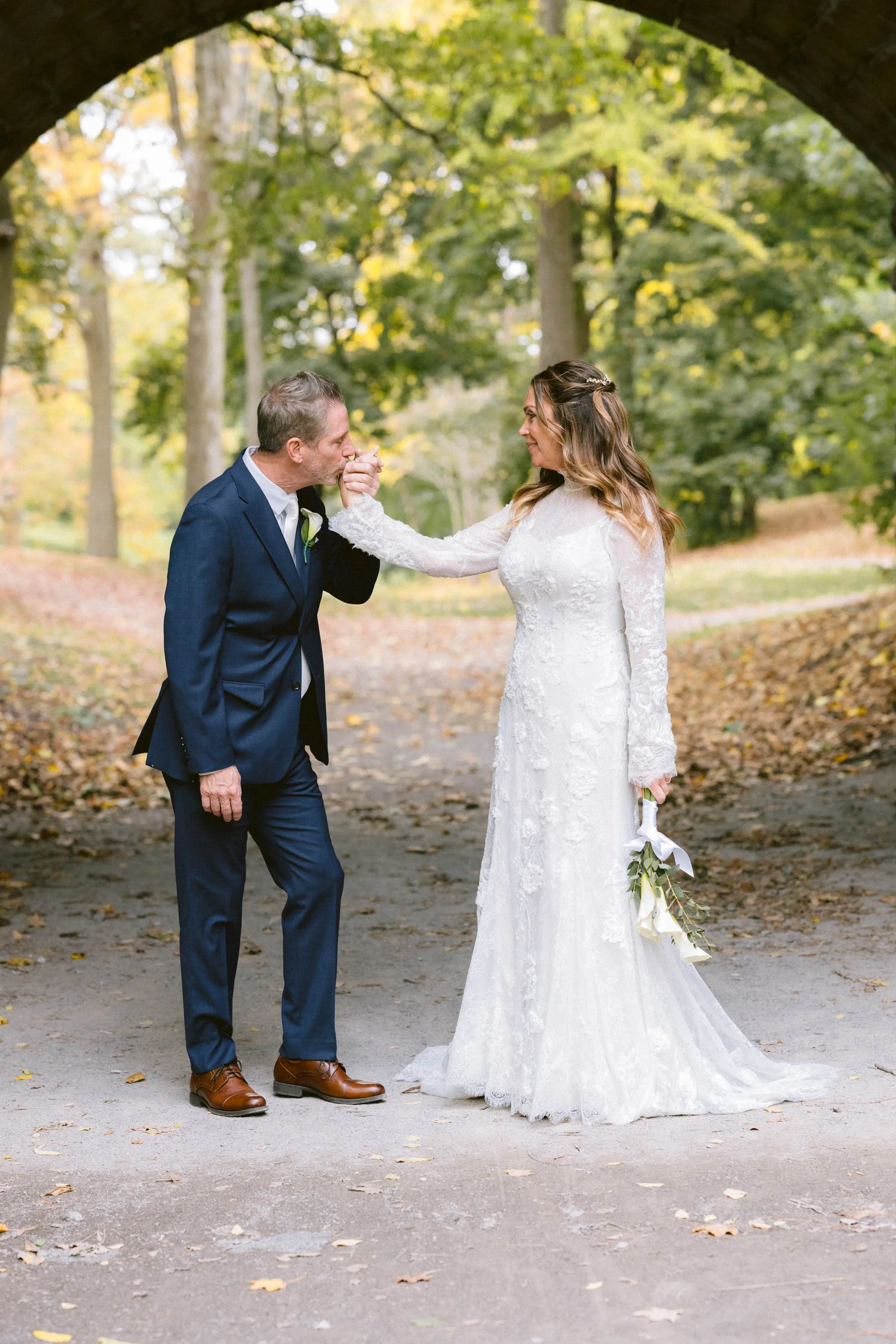 A bride in a white wedding dress kisses her father on the cheek under a bridge in a park with fall foliage.