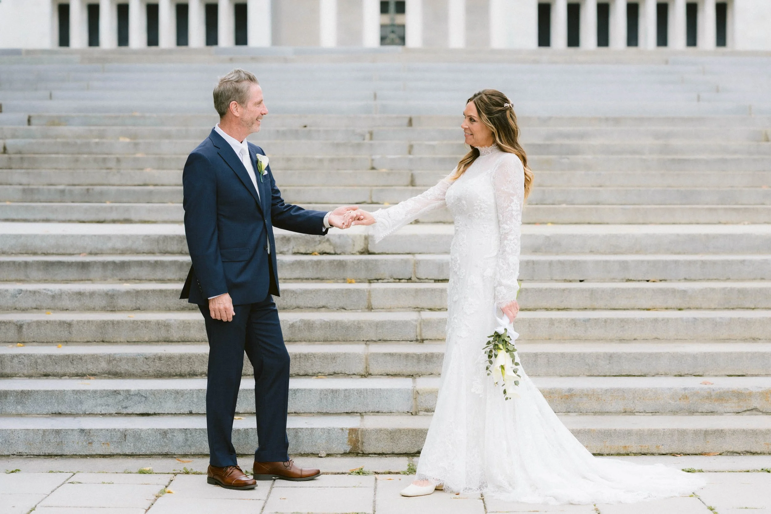 A bride and groom hold hands in front of a staircase, with the groom in a navy suit and the bride in a white lace wedding gown holding a bouquet.