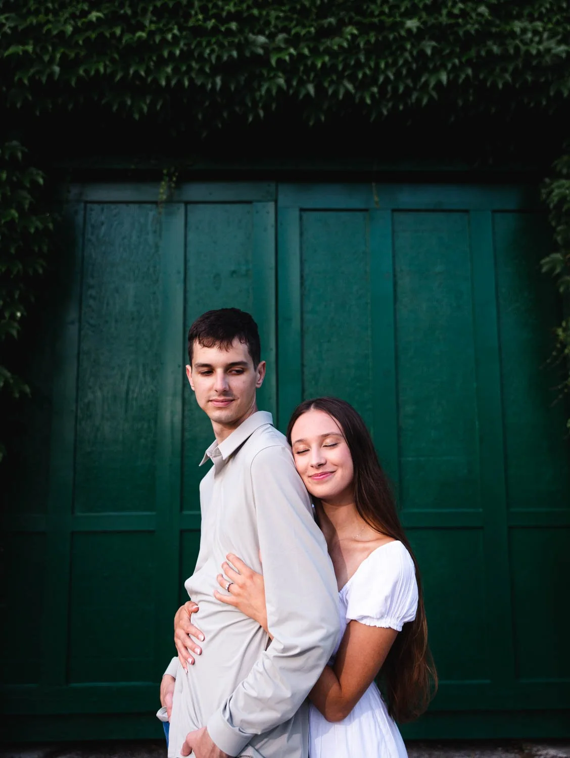 A young couple stands close together in front of a green wooden door, with the woman hugging the man from behind, both smiling softly.