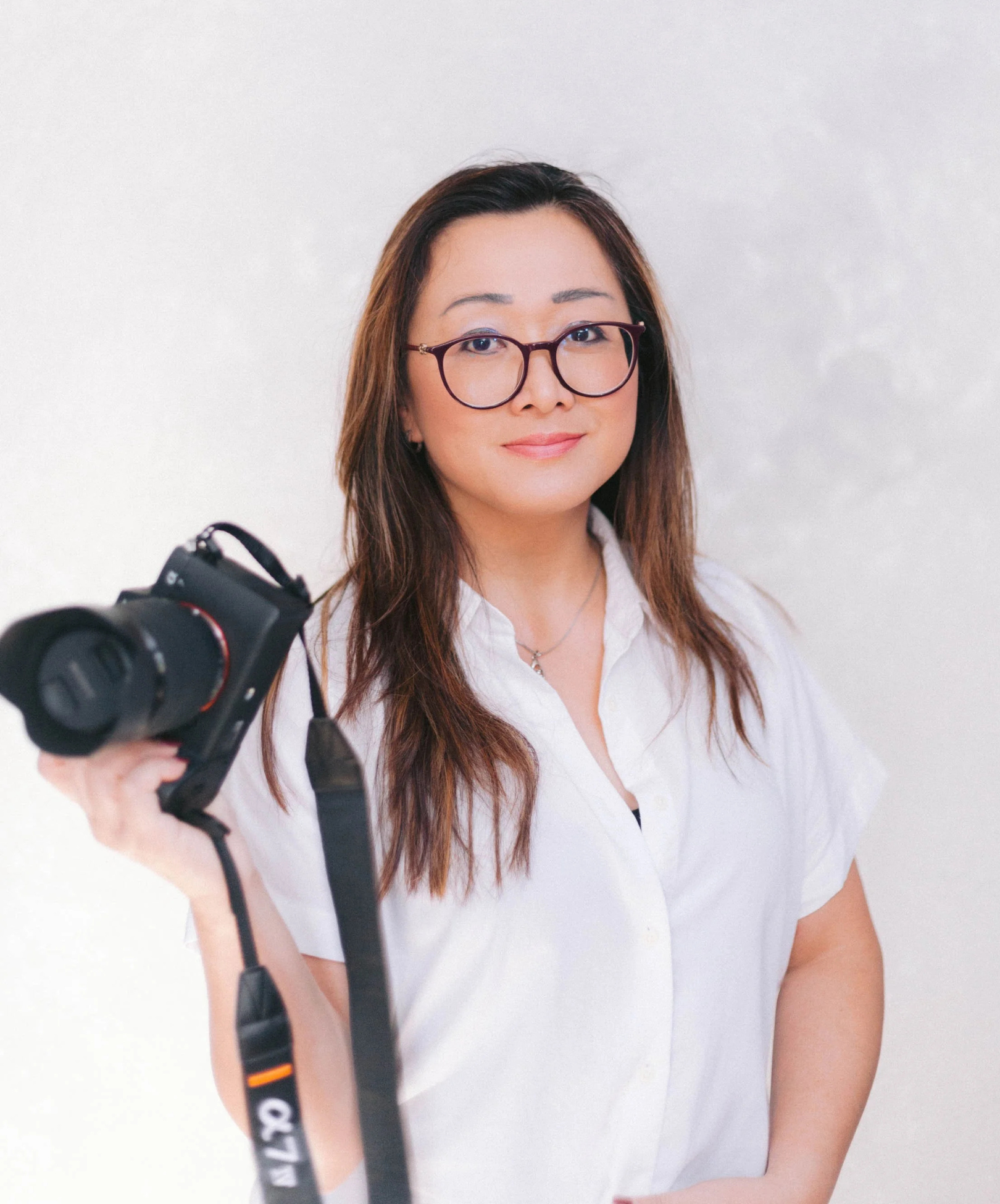 Woman with glasses holding a camera, wearing a white shirt, standing against a light-colored background.