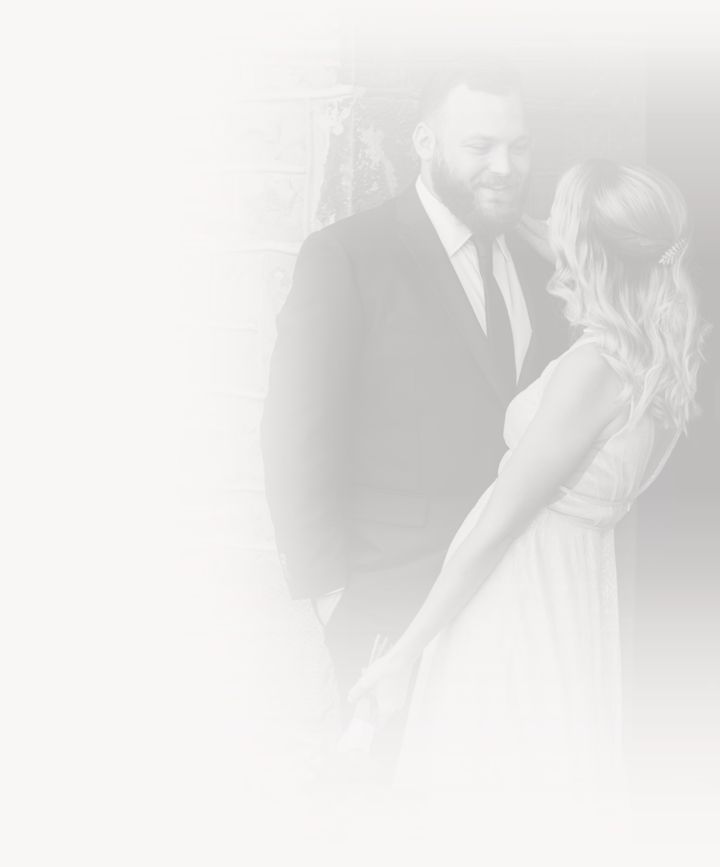 A man and woman dressed in formal attire standing close and smiling at each other outdoors near a brick wall.