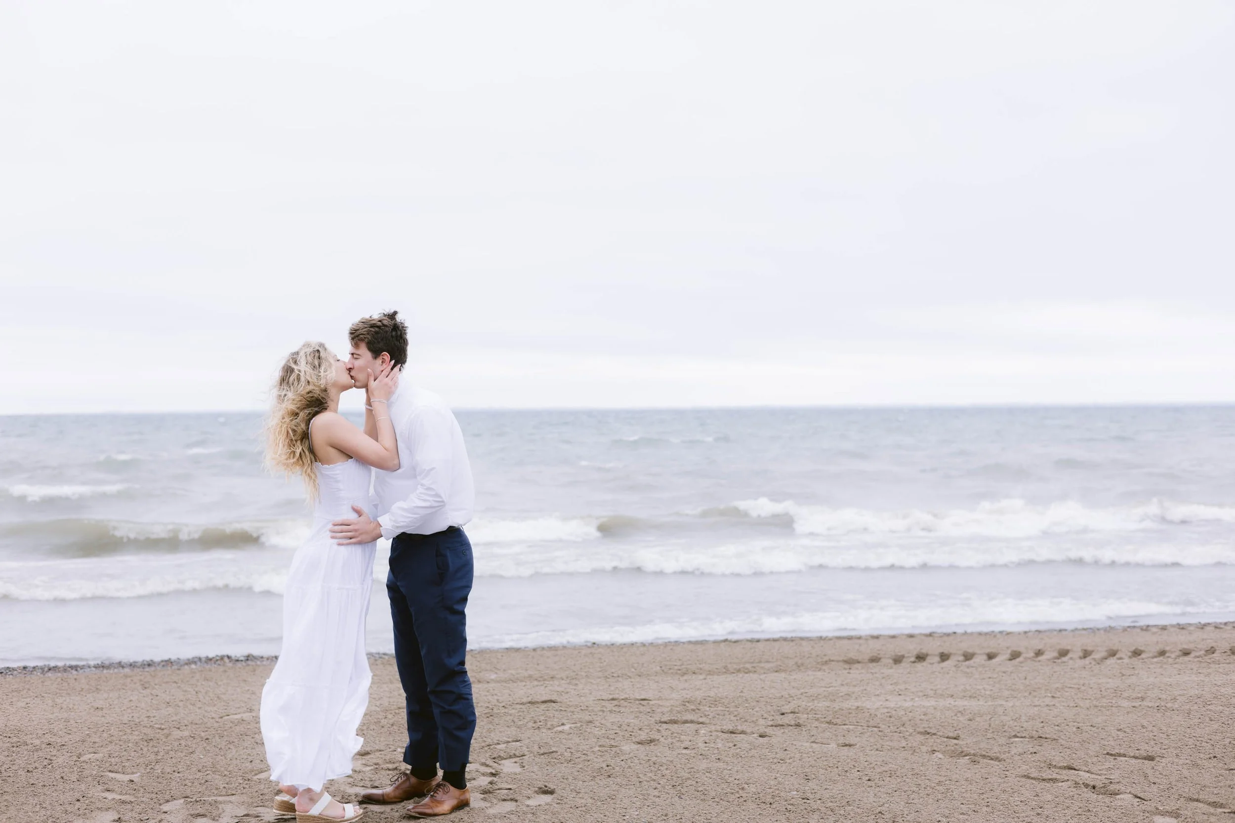 A couple embracing and kissing on a sandy beach near the ocean, overcast sky in the background.