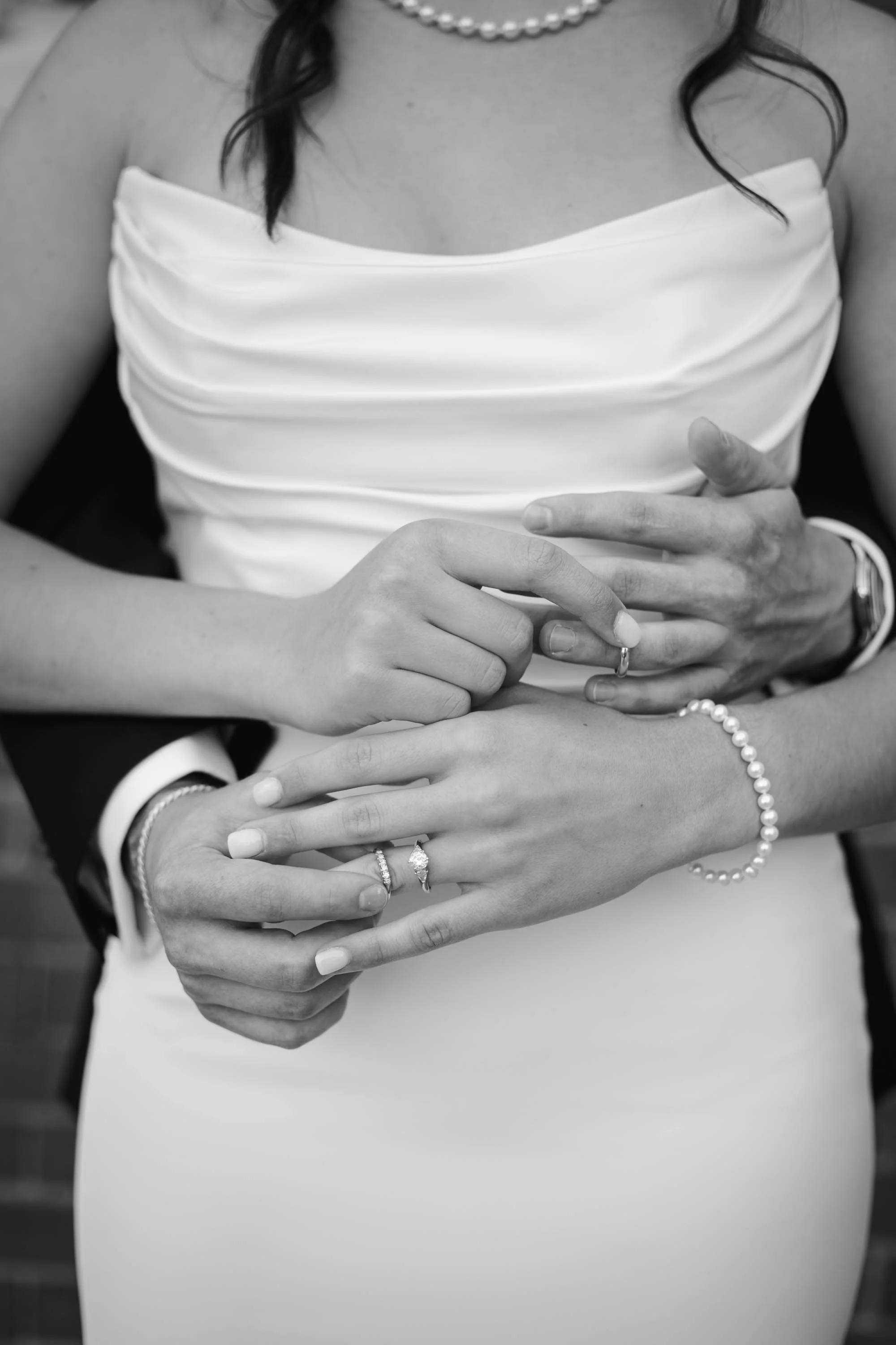 Black and white close-up of a couple's hands showing wedding rings, with the woman wearing a pearl bracelet and a pearl necklace, and the man wearing a watch, on a woman wearing a strapless dress.