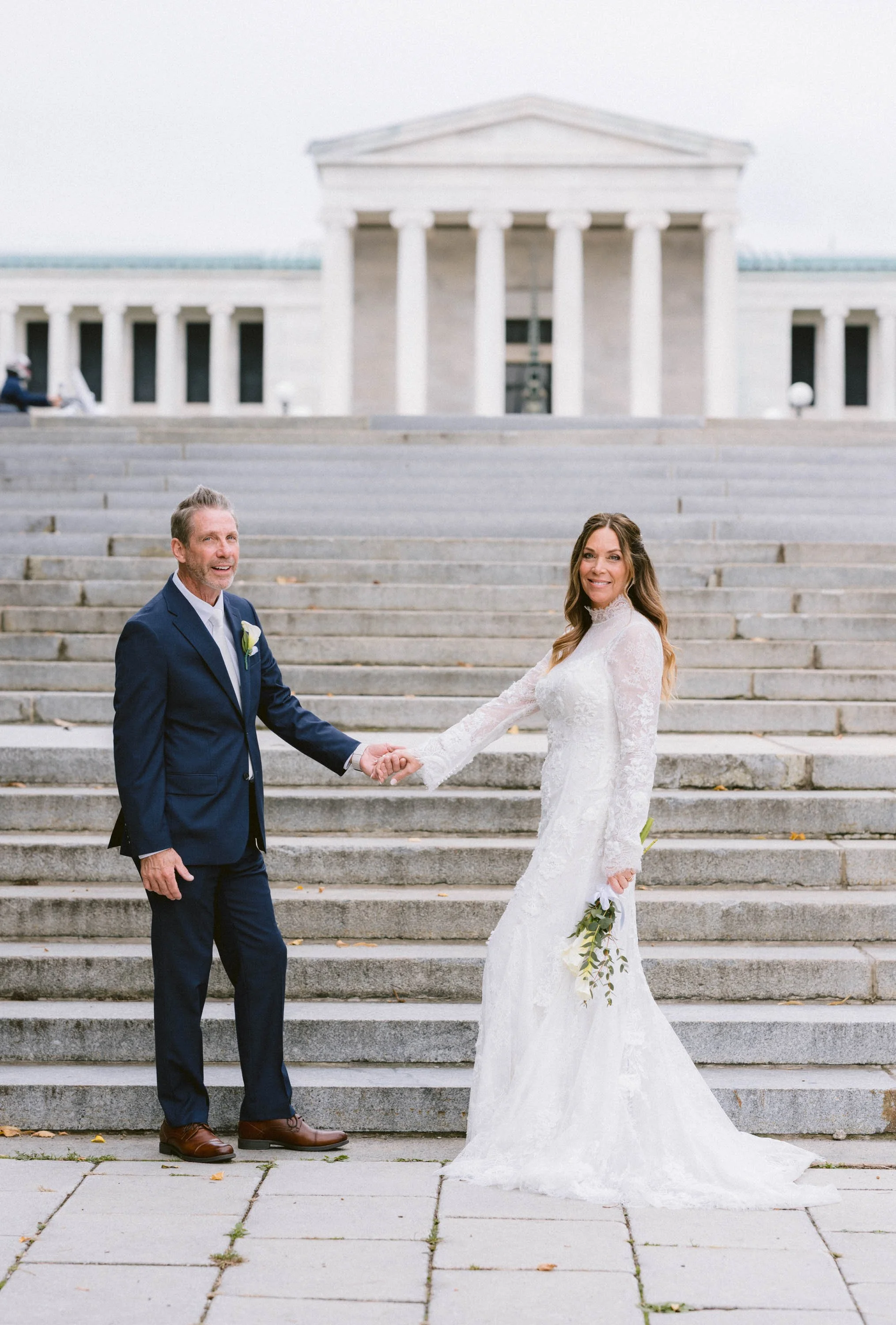 A bride in a white lace wedding dress and a man in a navy suit stand on steps in front of a neoclassical building, holding hands and smiling.
