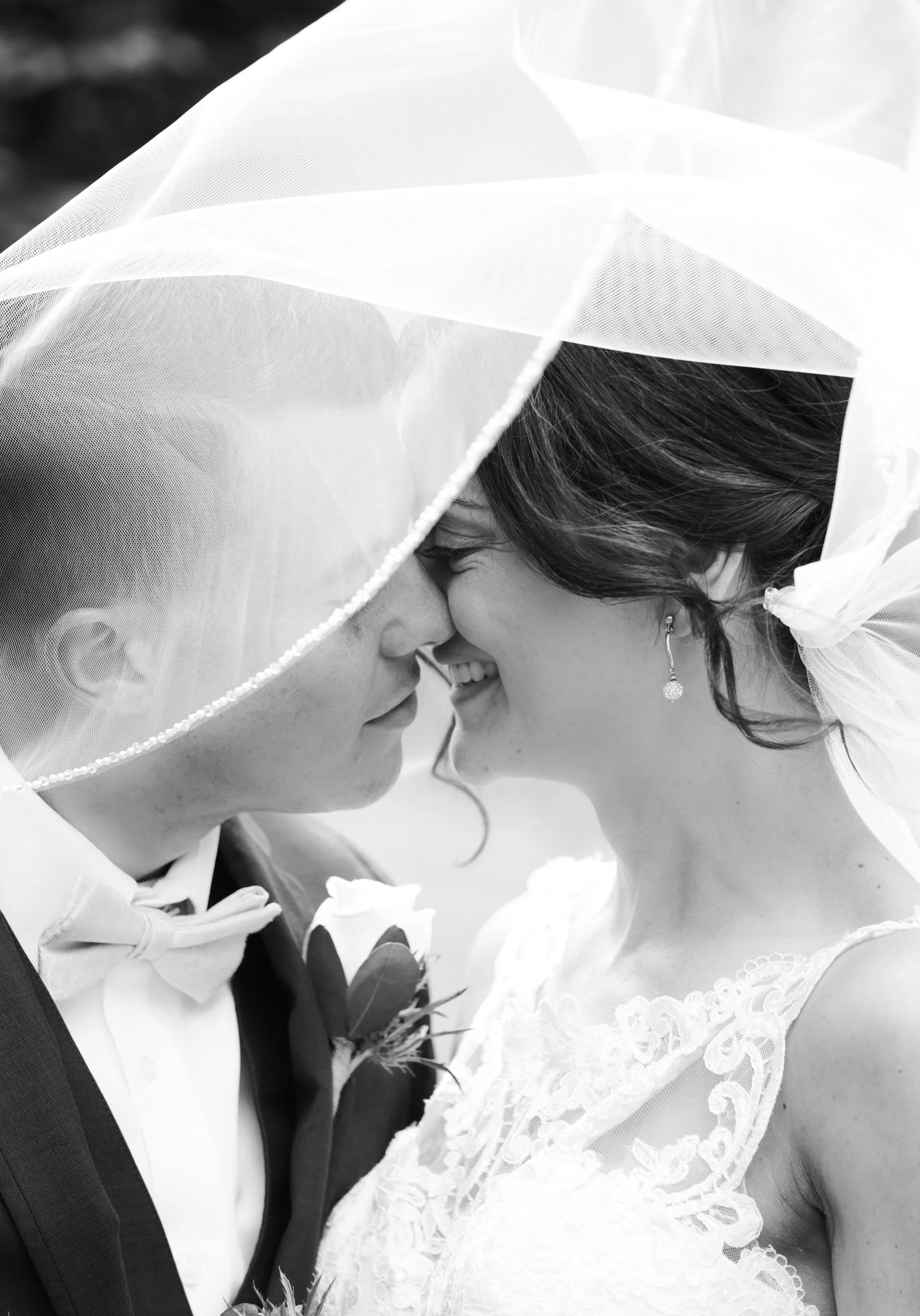 A black-and-white photo of a bride and groom with their foreheads touching, smiling, under a veil on their wedding day.