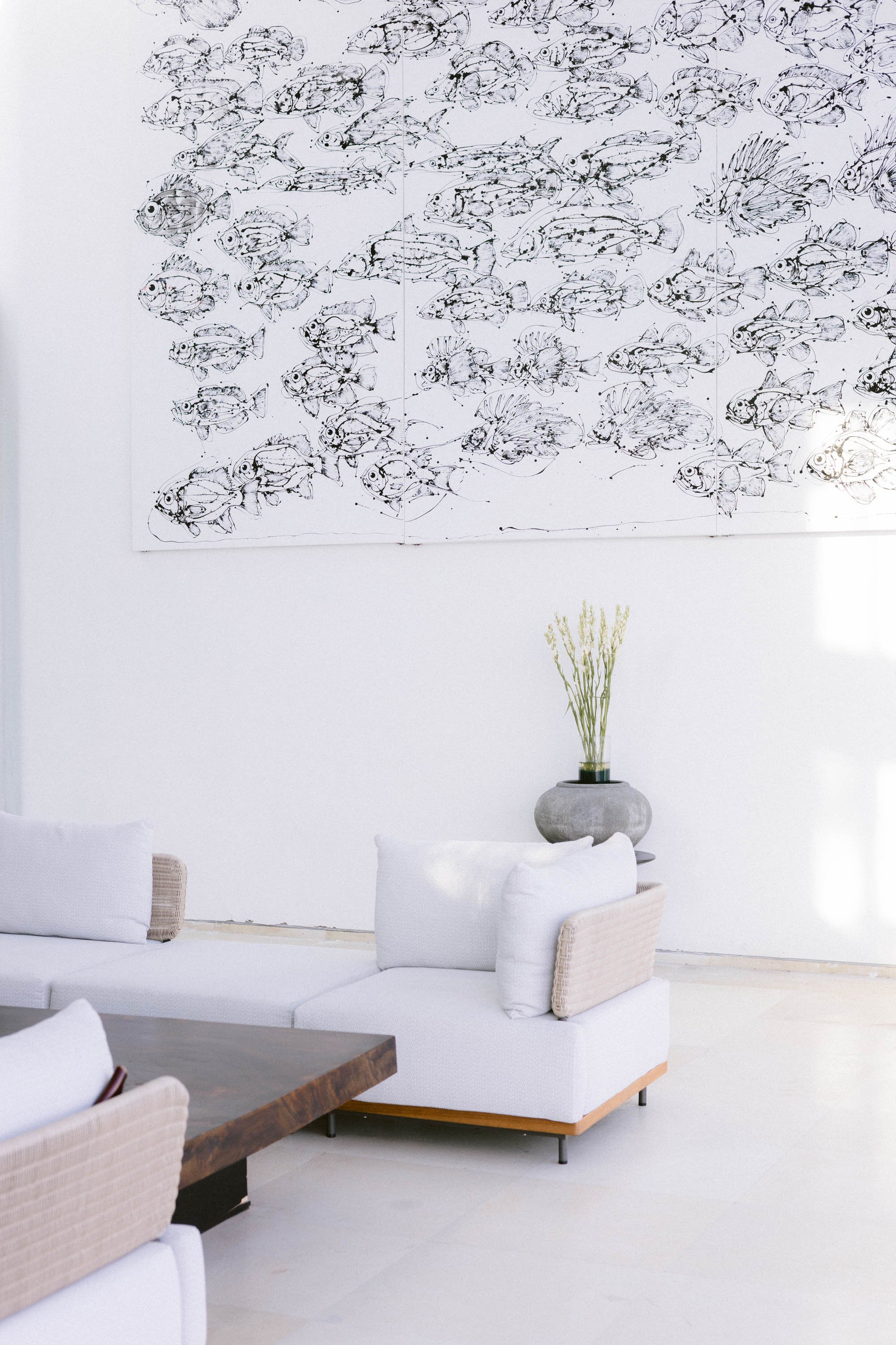 Minimalist living room with a white sofa, pillows, a wooden table, a decorative vase with green tall grass, and wall art of pencil sketches of fish.