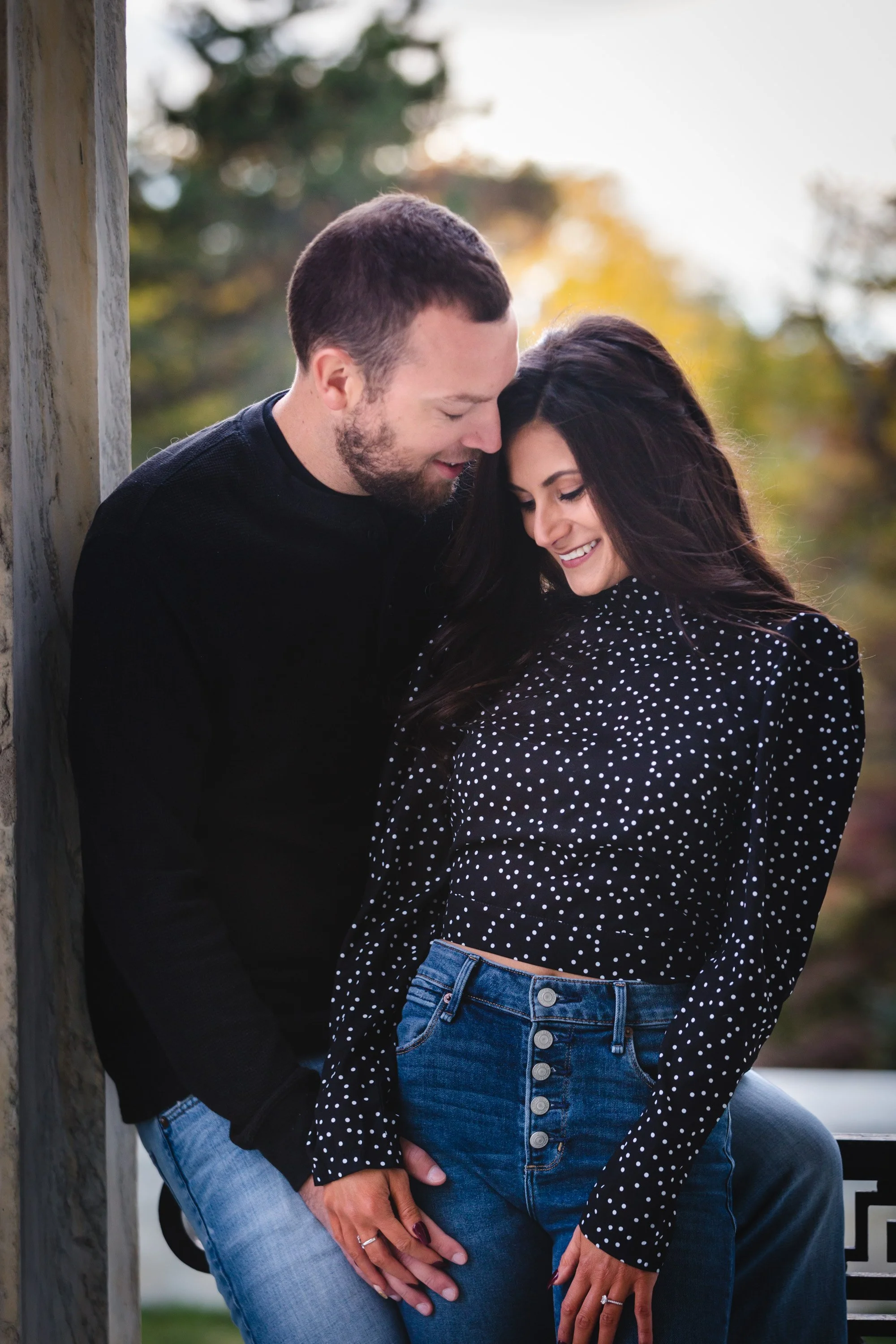 A couple sharing an intimate moment outdoors during autumn, leaning against a wooden pillar, smiling, with trees in the background.