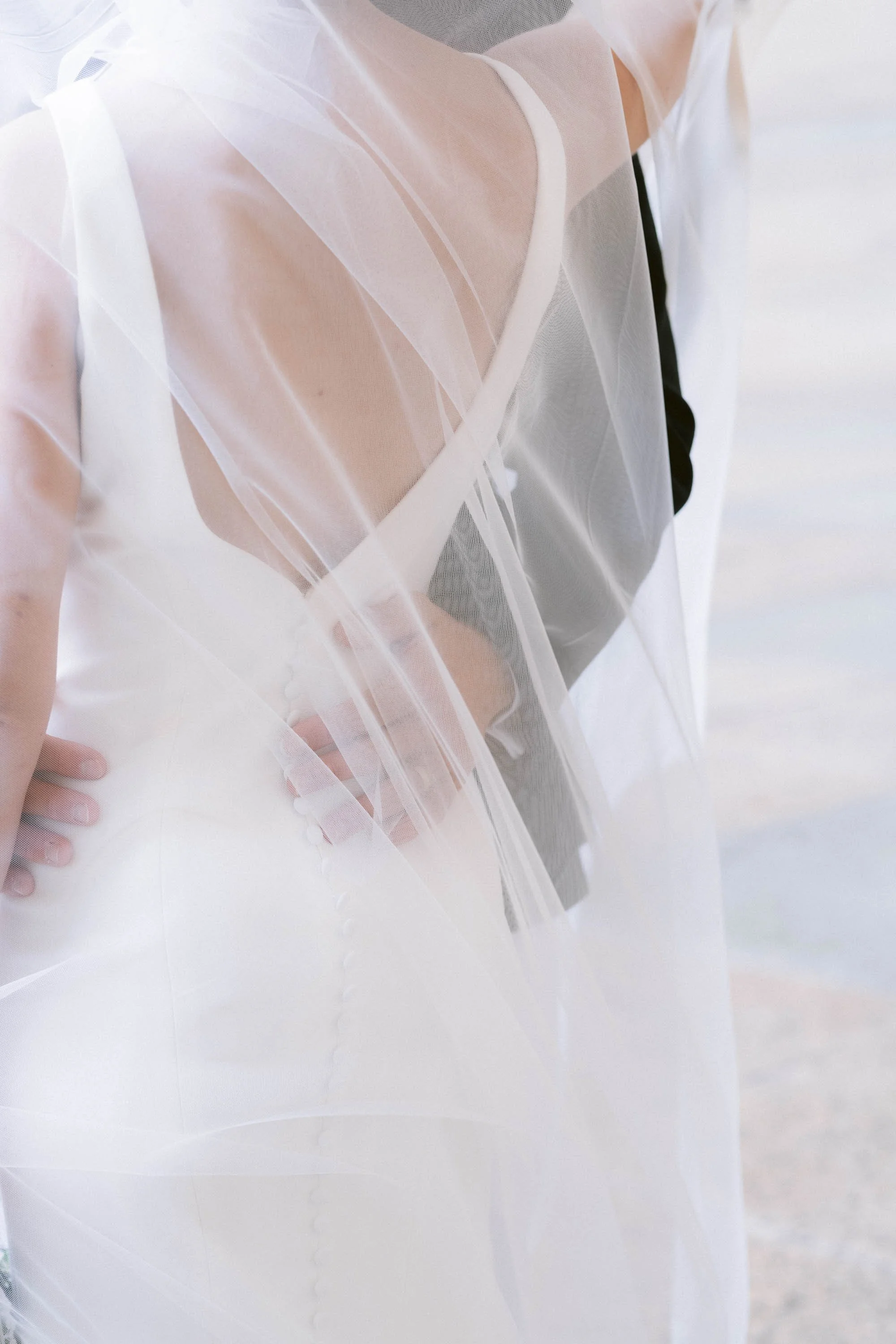 Close-up of a bride in a wedding dress with sheer fabric, standing by a body of water.