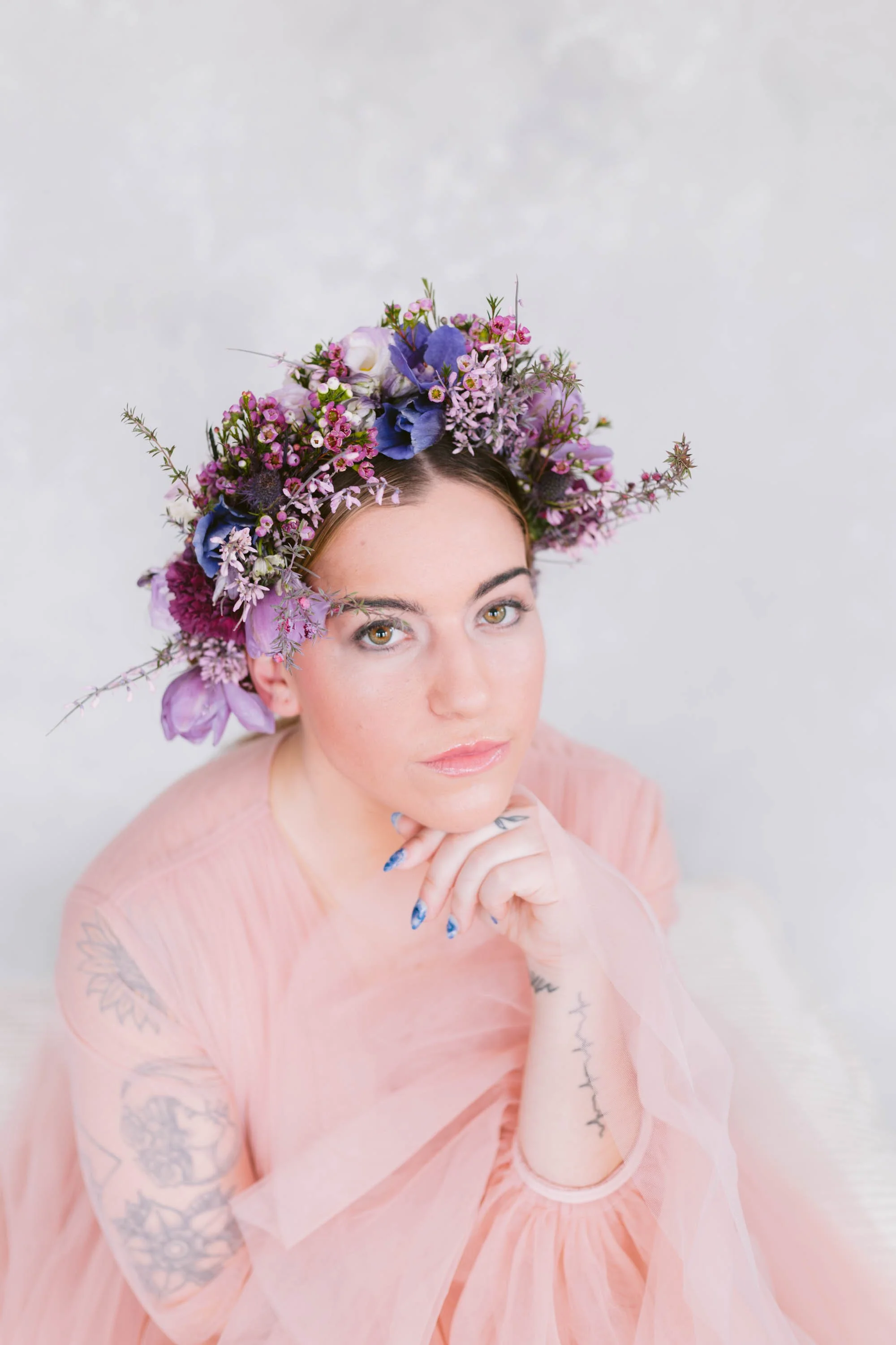 Portrait of a woman with a floral crown made of purple, pink, and blue flowers, wearing a light pink sheer dress, with tattoos on her arms, resting her chin on her hand.