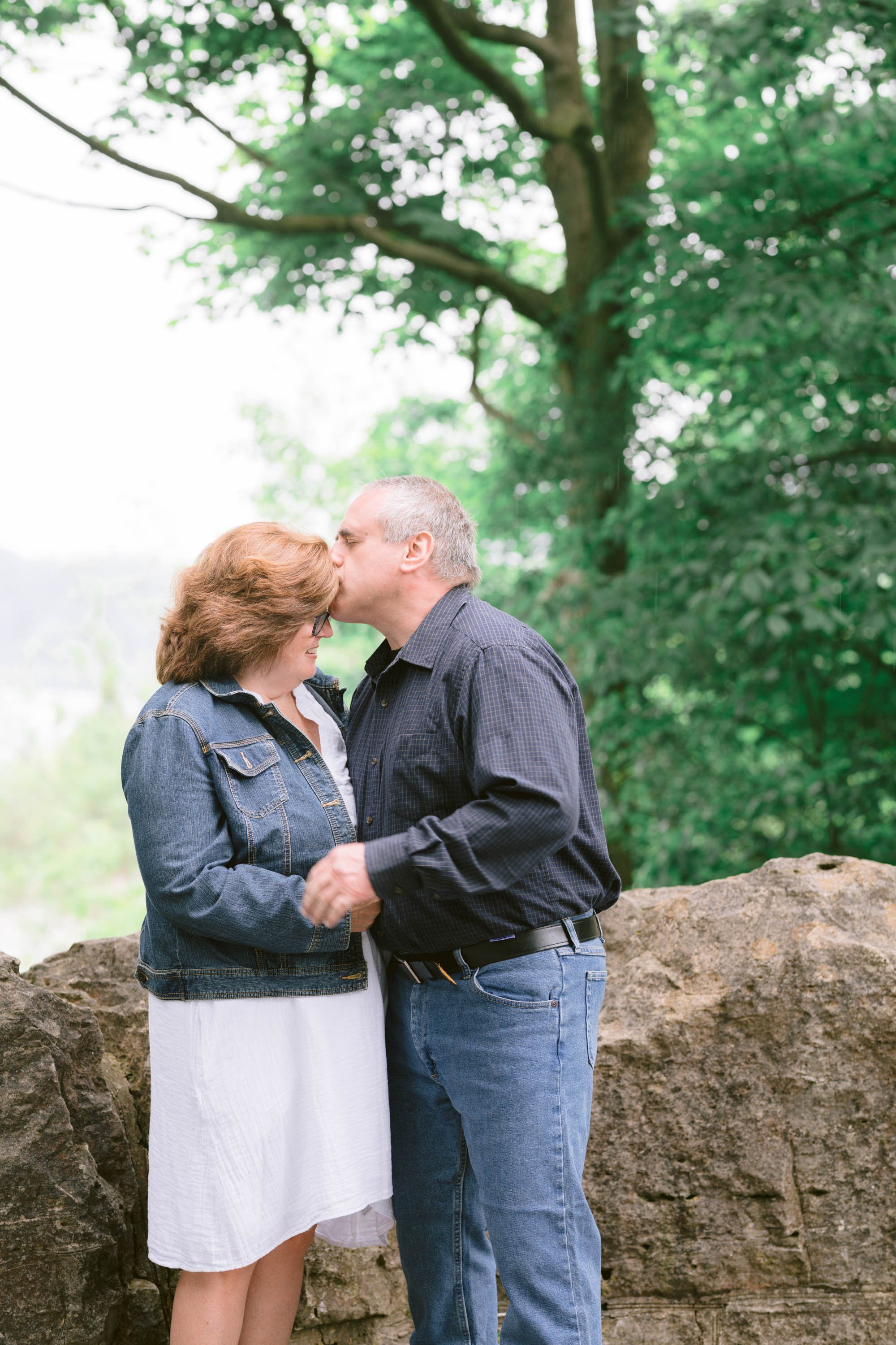 A couple stands close together, with the man kissing the woman's forehead, in an outdoor setting with green trees and a rocky background.