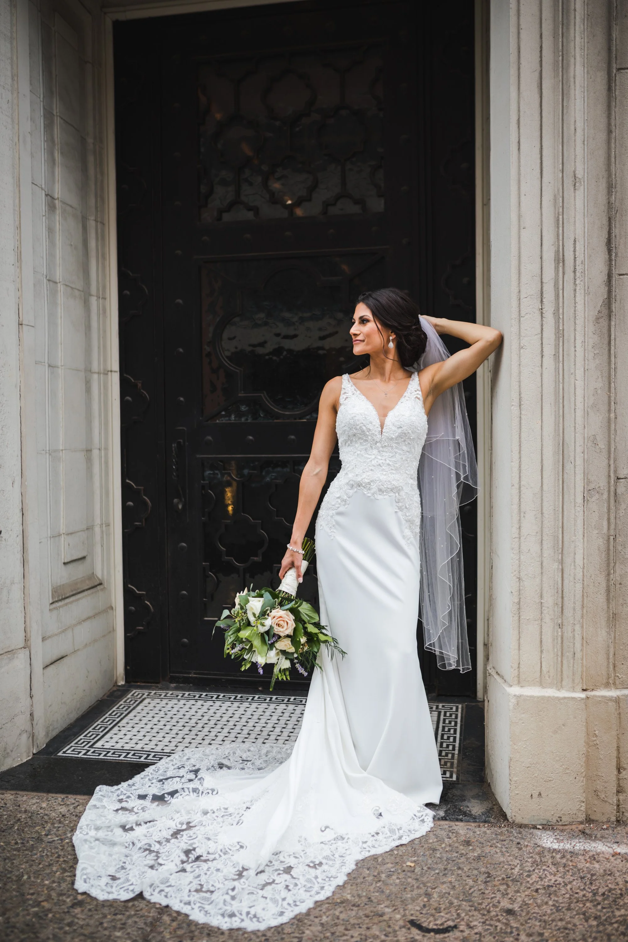 A bride in a white wedding dress holding a bouquet, standing in front of a black ornate door on a city street.