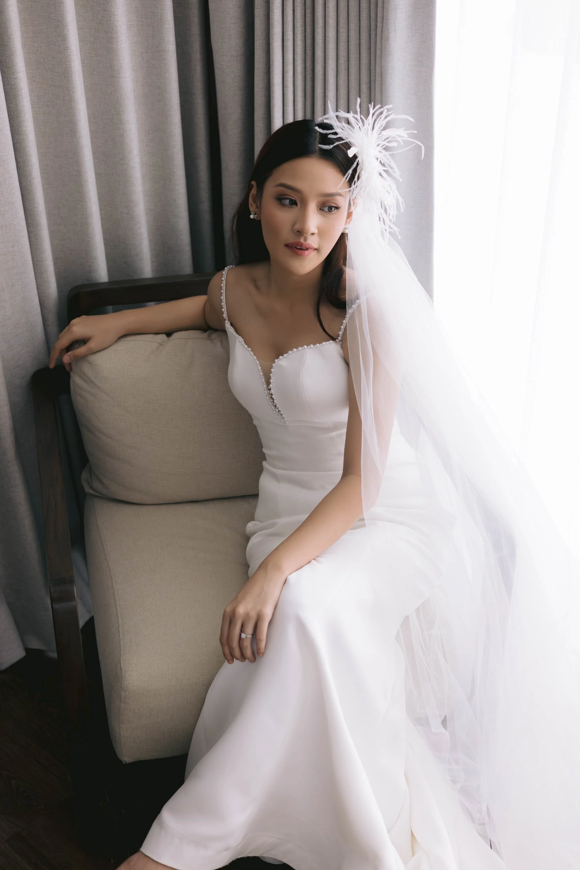 A bride in a white wedding dress sitting on a beige sofa by a window with gray curtains.