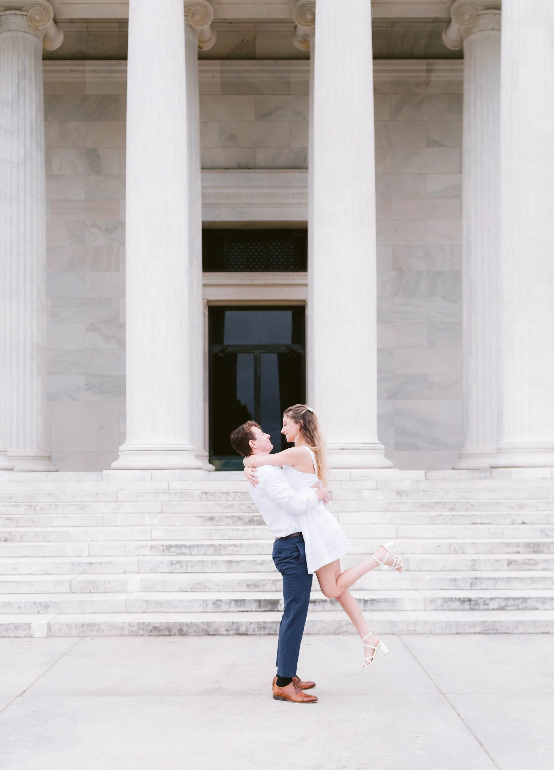 A man lifting a woman in a white dress in front of large classical columns and steps of a building.