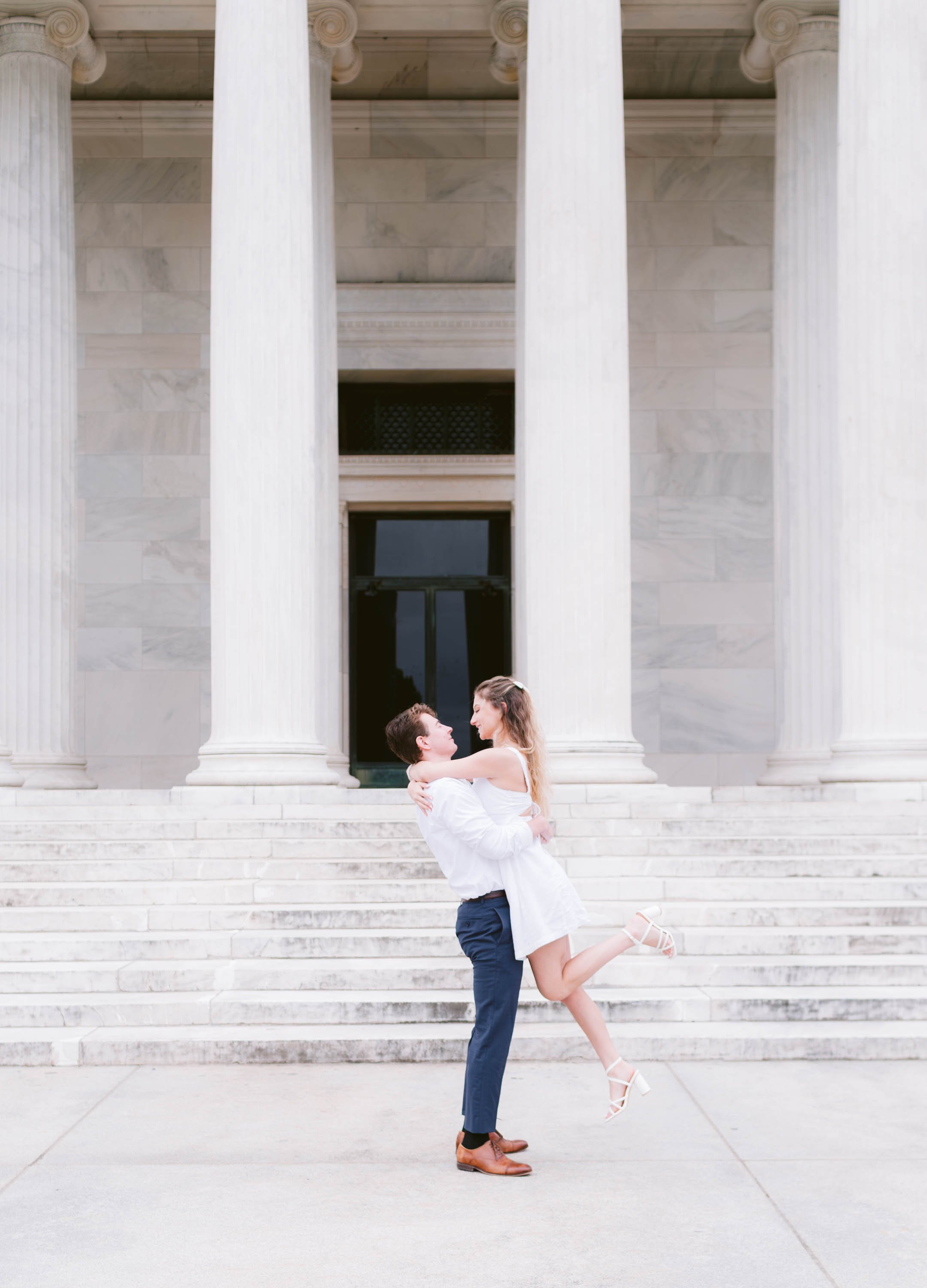 A man lifting a woman in a white dress in front of large classical columns and steps of a building.