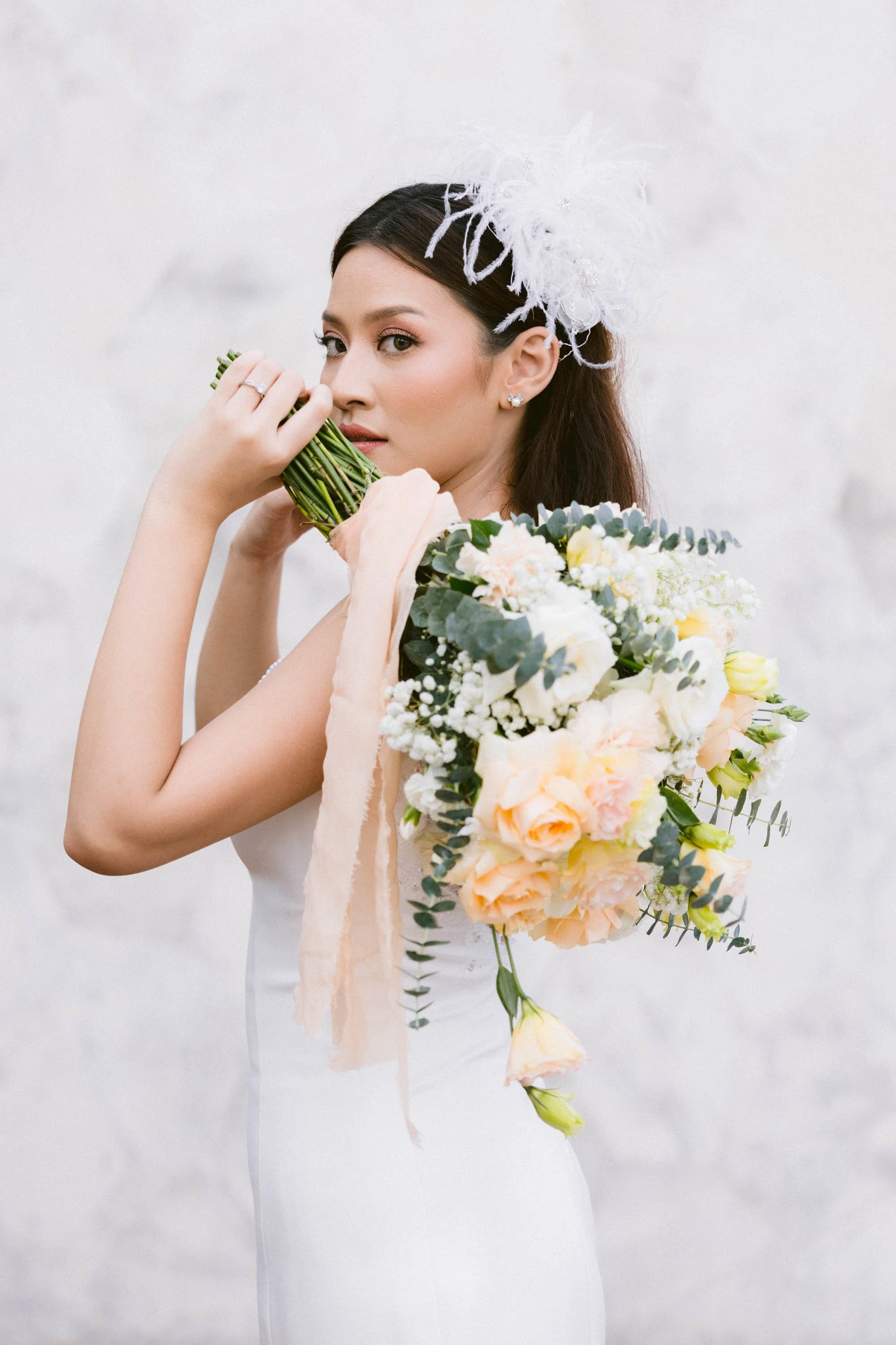 A woman in a wedding dress holding a large bouquet of flowers and a bunch of green herbs, with a white headpiece, standing against a white wall.