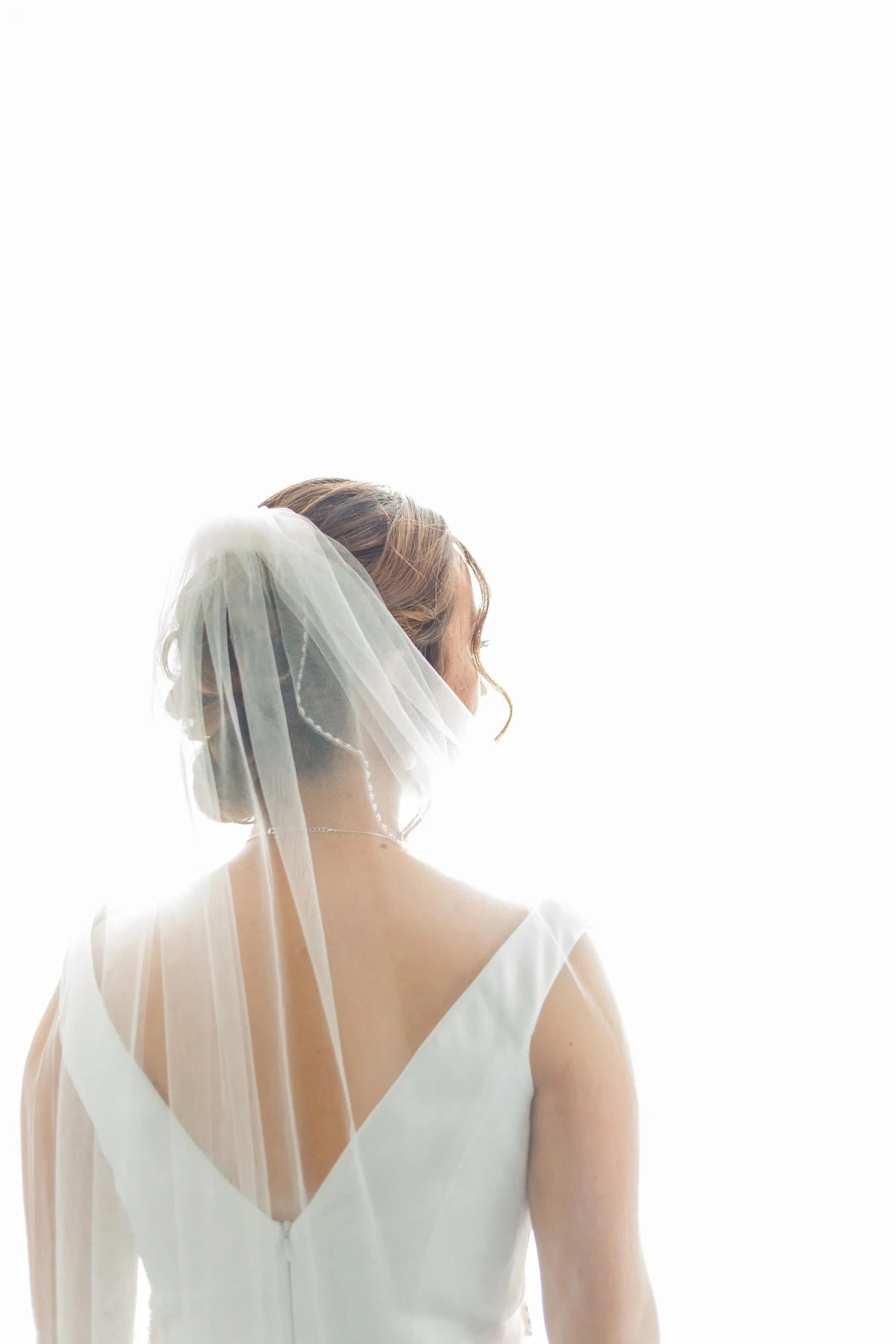 A bride with a veil and wedding dress, her face turned away, against a bright, white background.
