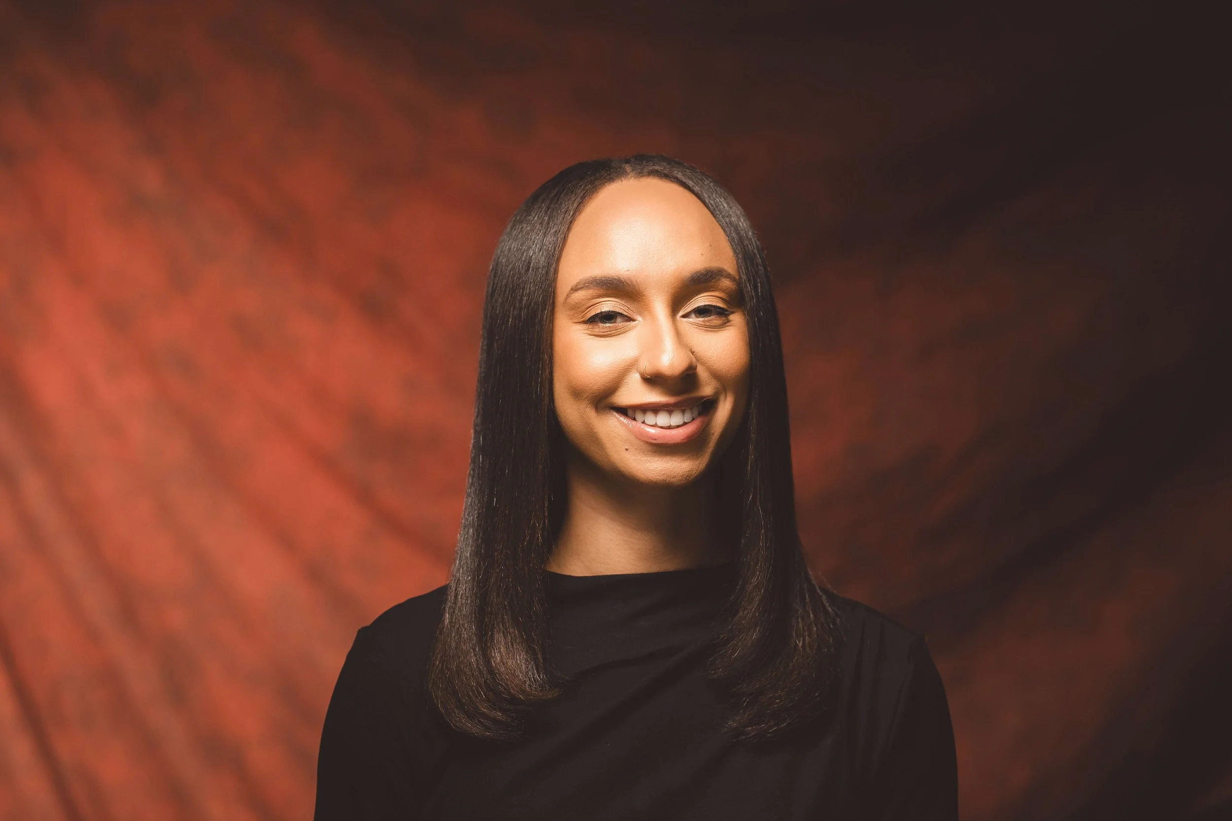 A young woman with long dark hair, wearing a black top, smiling against a red and black blurred background.