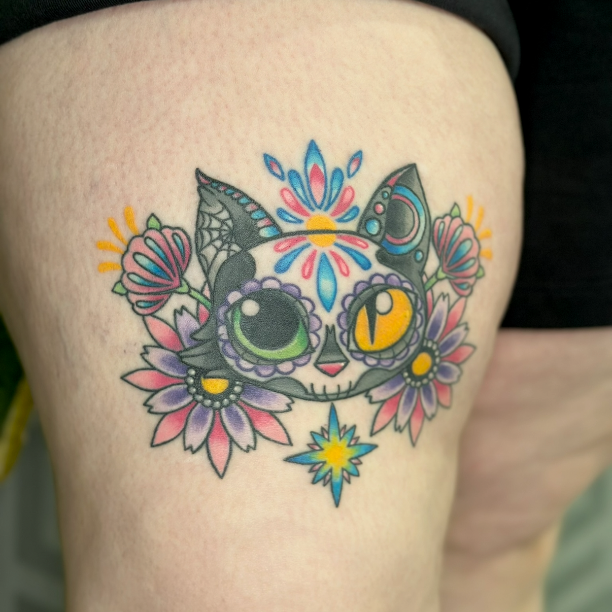 Colorful tattoo of a sugar skull cat with floral decorations and ornaments, featuring bright colors like pink, purple, yellow, blue, and green.