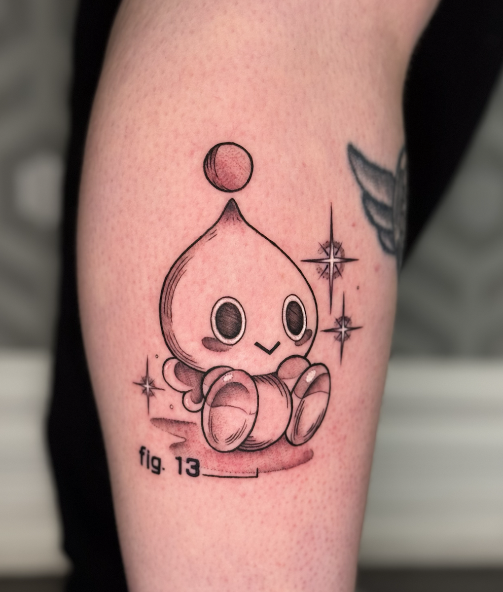 Vintage encyclopedia style tattoo of a cute, plump Chao from Sonic the Hedgehog with large eyes, sitting with its legs crossed, and a small circle above its head, with decorative stars around it and the label 'fig. 13' underneath.