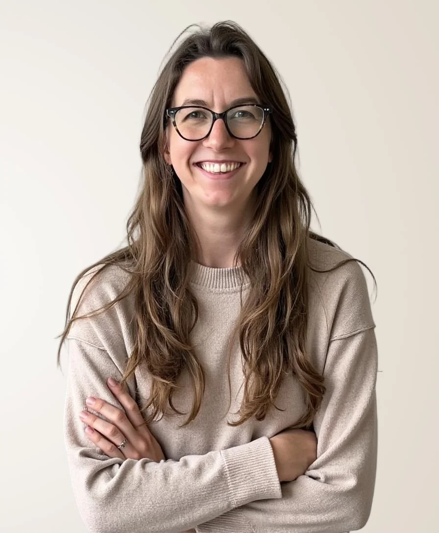 Tayler Kellim, a postpartum doula, with long wavy brown hair and black glasses smiling and crossing her arms in front of a plain white wall.