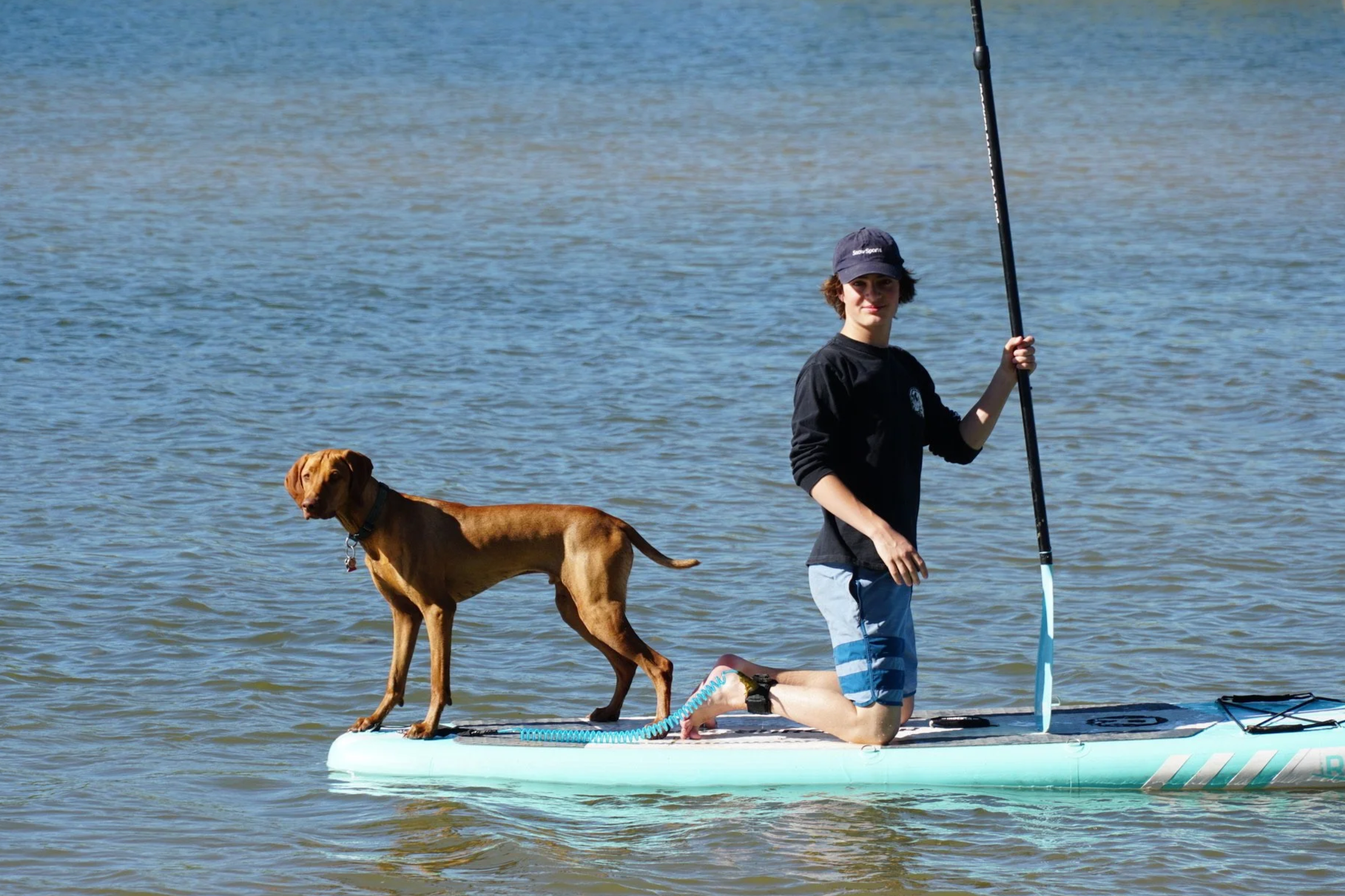 A young man kneeling on a paddleboard holding a paddle, with a brown dog standing beside him on the paddleboard, in a body of water.