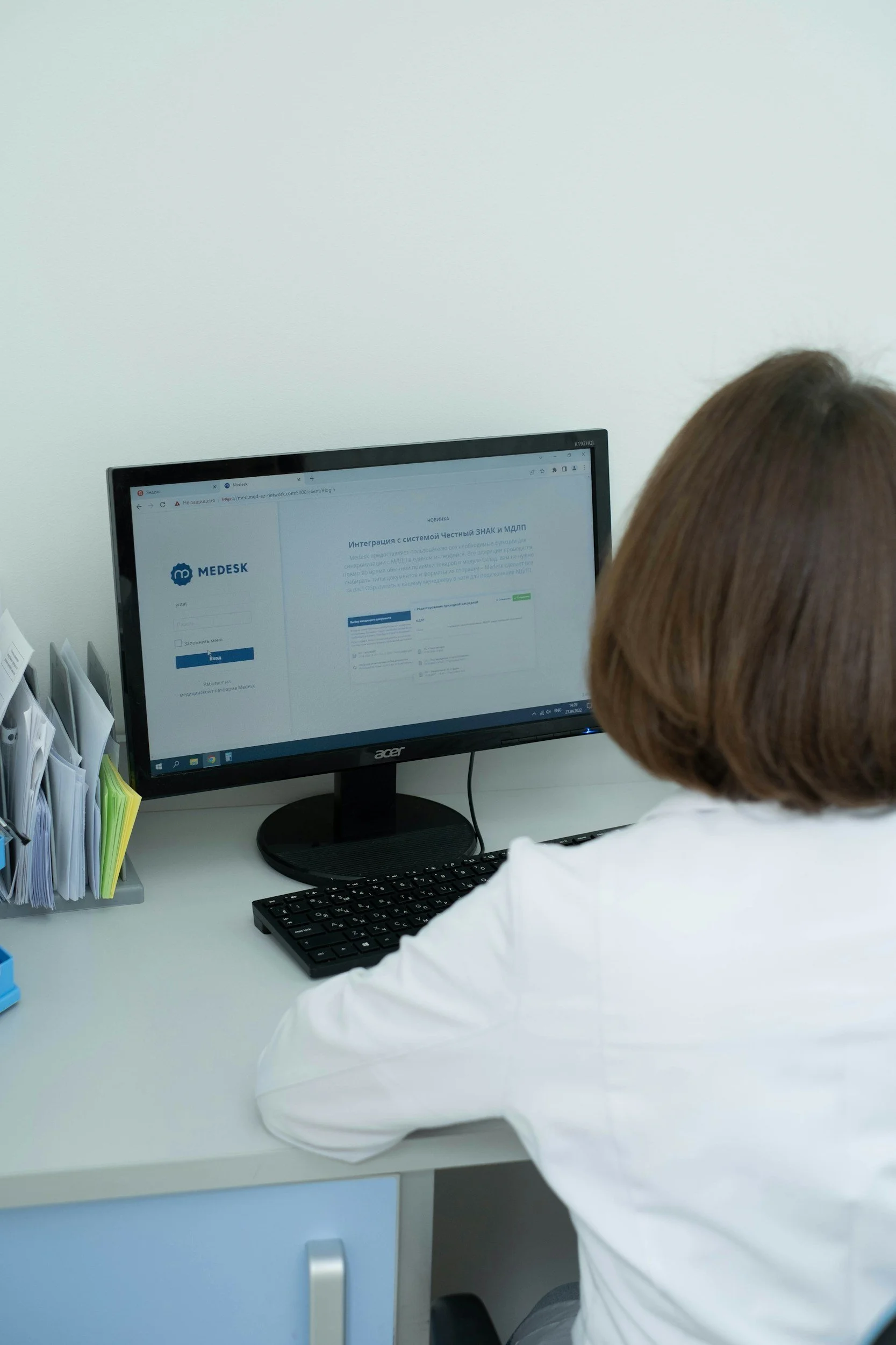 A woman in a white coat sitting at a desk, working on a computer with a webpage open that shows the MEDISK logo and text in Russian.