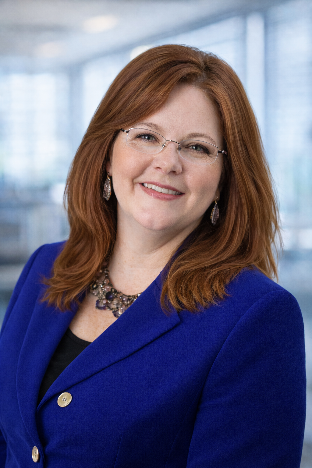 A woman with red hair, glasses, and earrings, wearing a blue blazer and a necklace, smiling in a professional setting with a blurred modern interior background.
