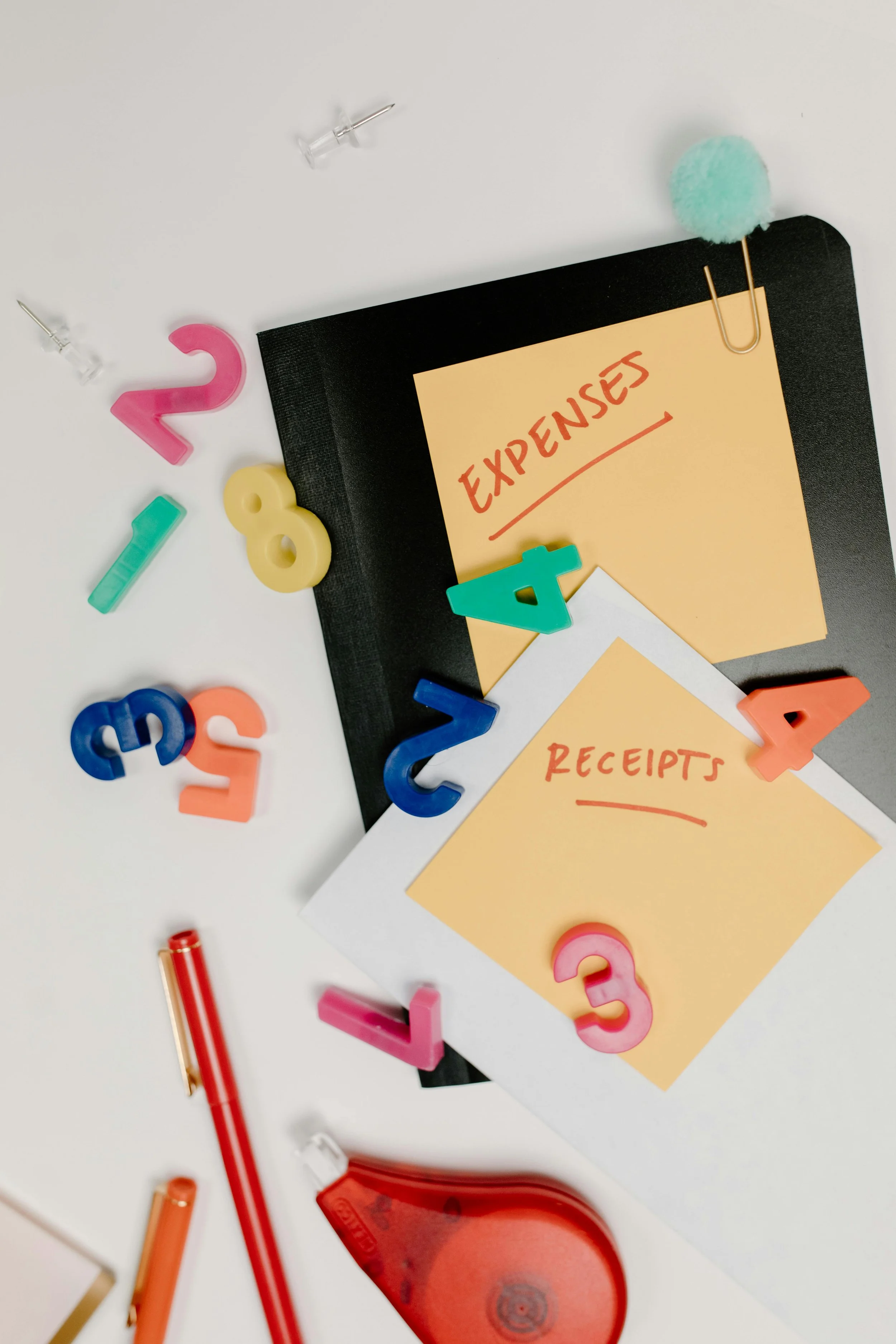 A desk with a black binder, two notes labeled 'Expenses' and 'Receipts,' colorful magnetic numbers, push pins, a red correction tape, and a stapler.