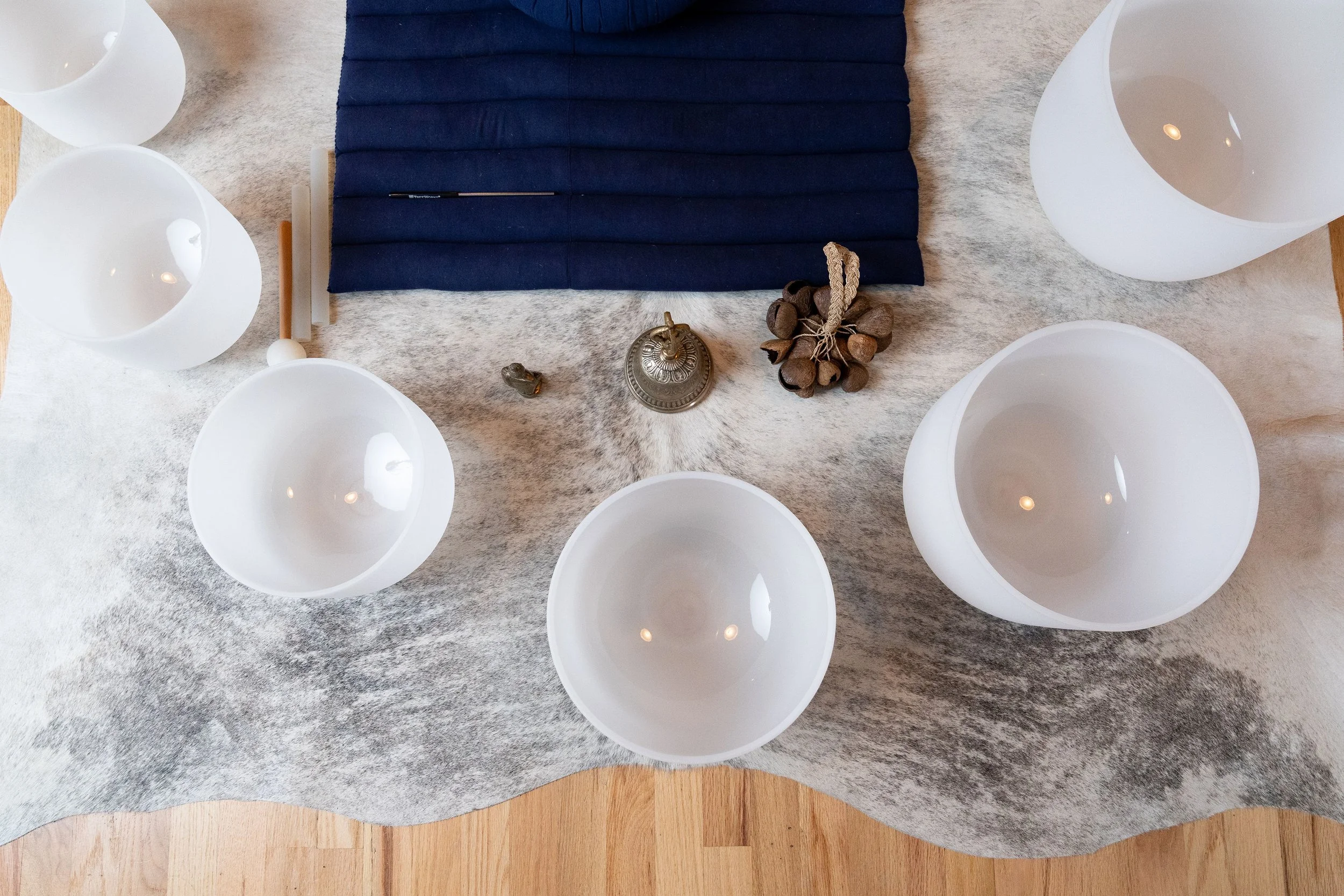 A table setting with five white bowls, a dark blue cloth, and decorative objects including a small brass bell, a bundle of dried seed pods, and a small figure, arranged on a textured gray table runner over a wood surface.