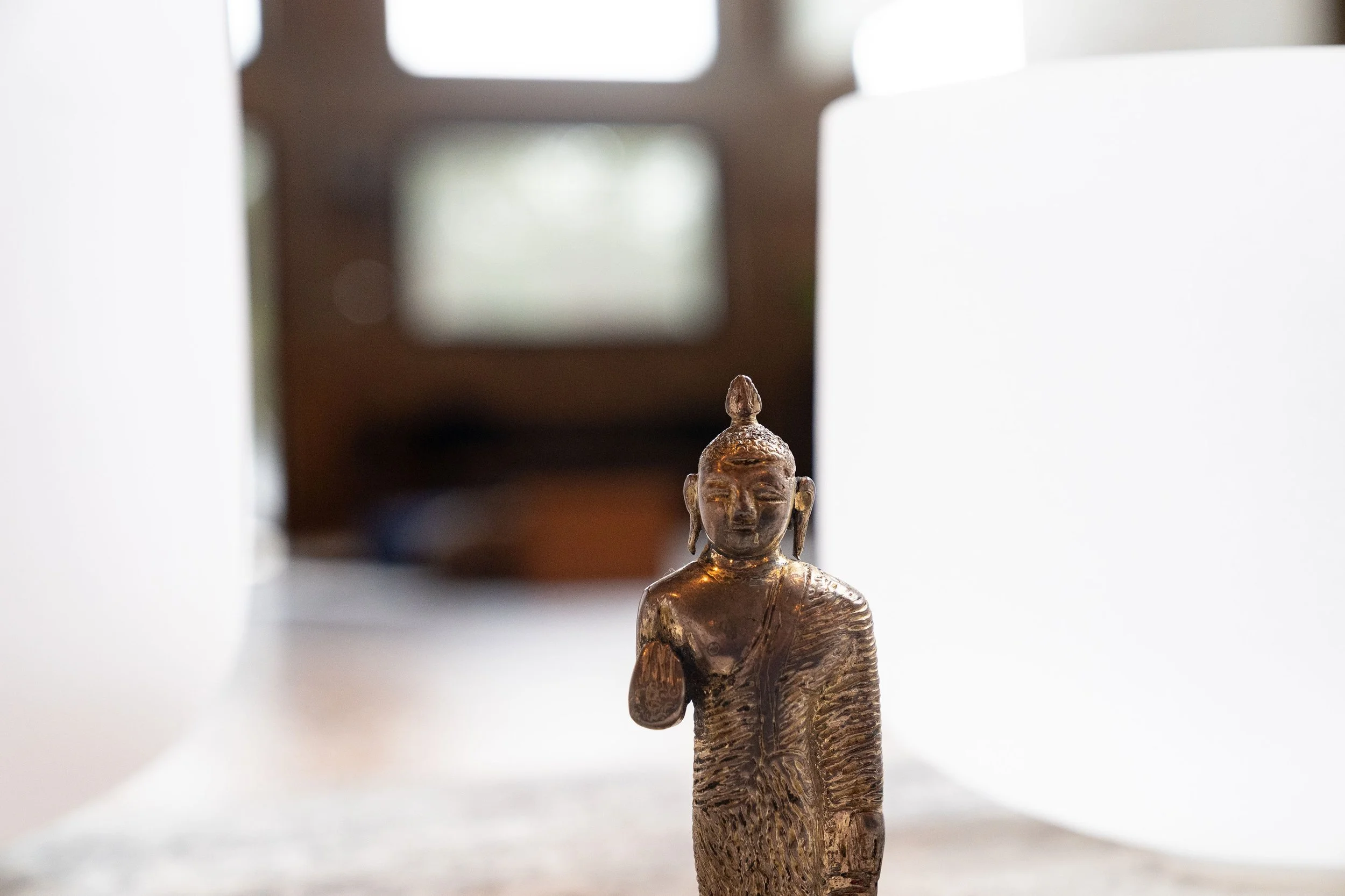 A small bronze Buddha statue on a table with white curtains on either side and a blurred wooden window or mirror in the background.