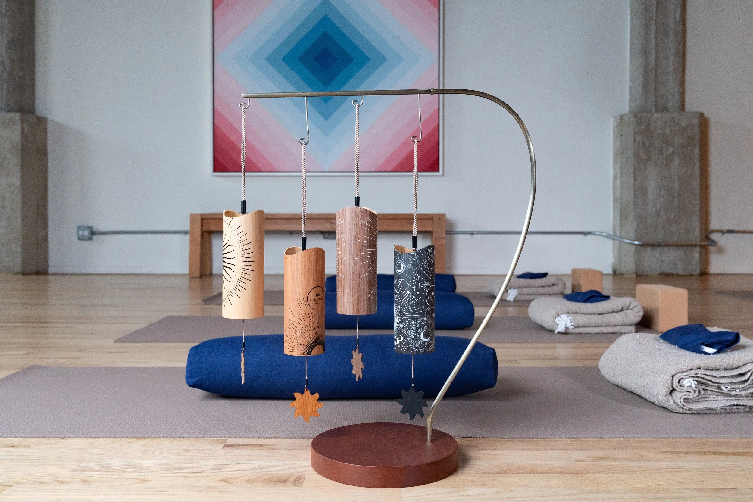 A yoga studio with mats, blankets, and blocks arranged on a wooden floor. A decorative chime with four hanging wooden cylinders and star-shaped ornaments is in the foreground. An abstract geometric artwork is on the wall in the background.