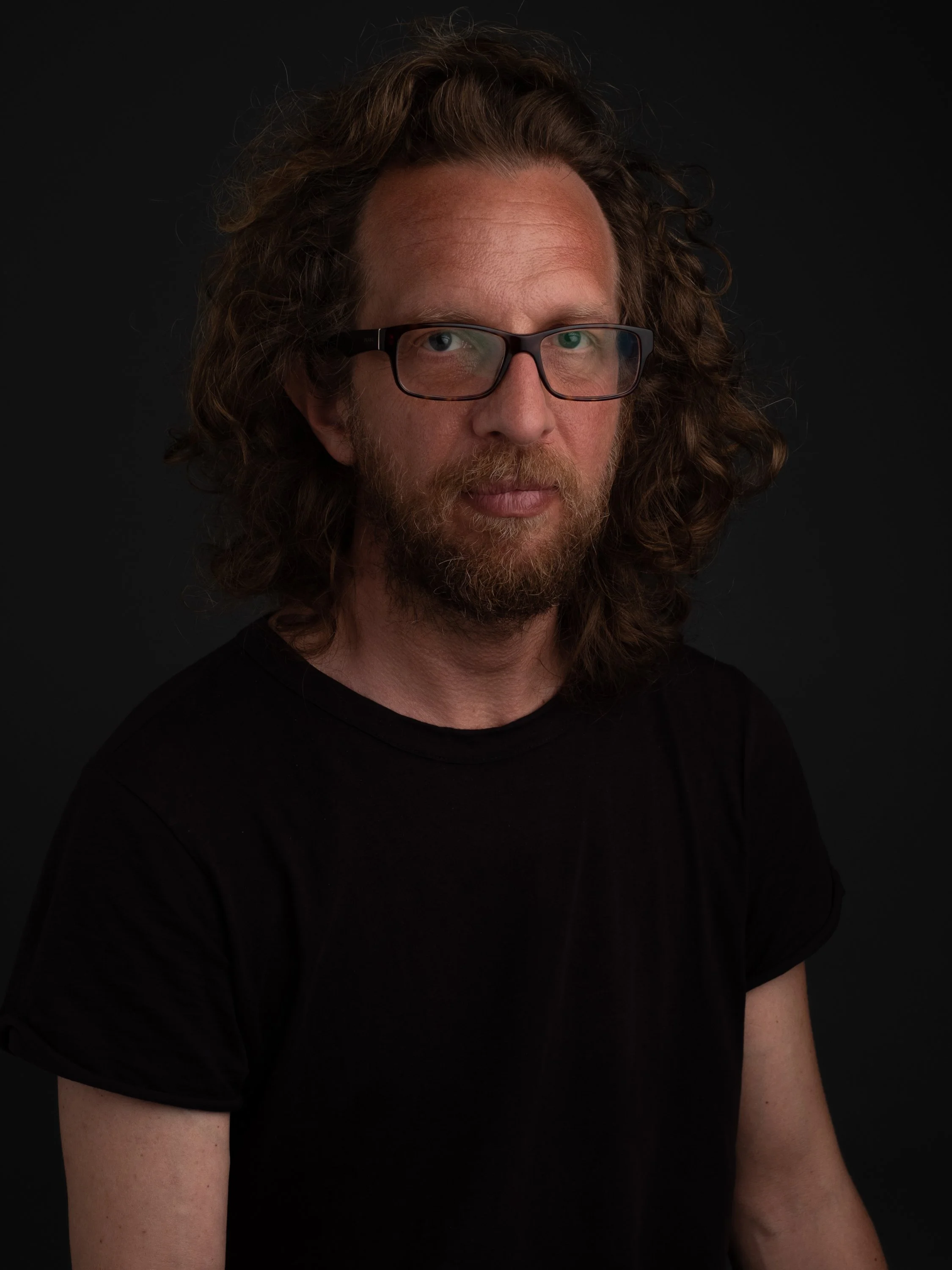 Retrato de un hombre con cabello rizado, barba, gafas y camiseta negra, en fondo oscuro.