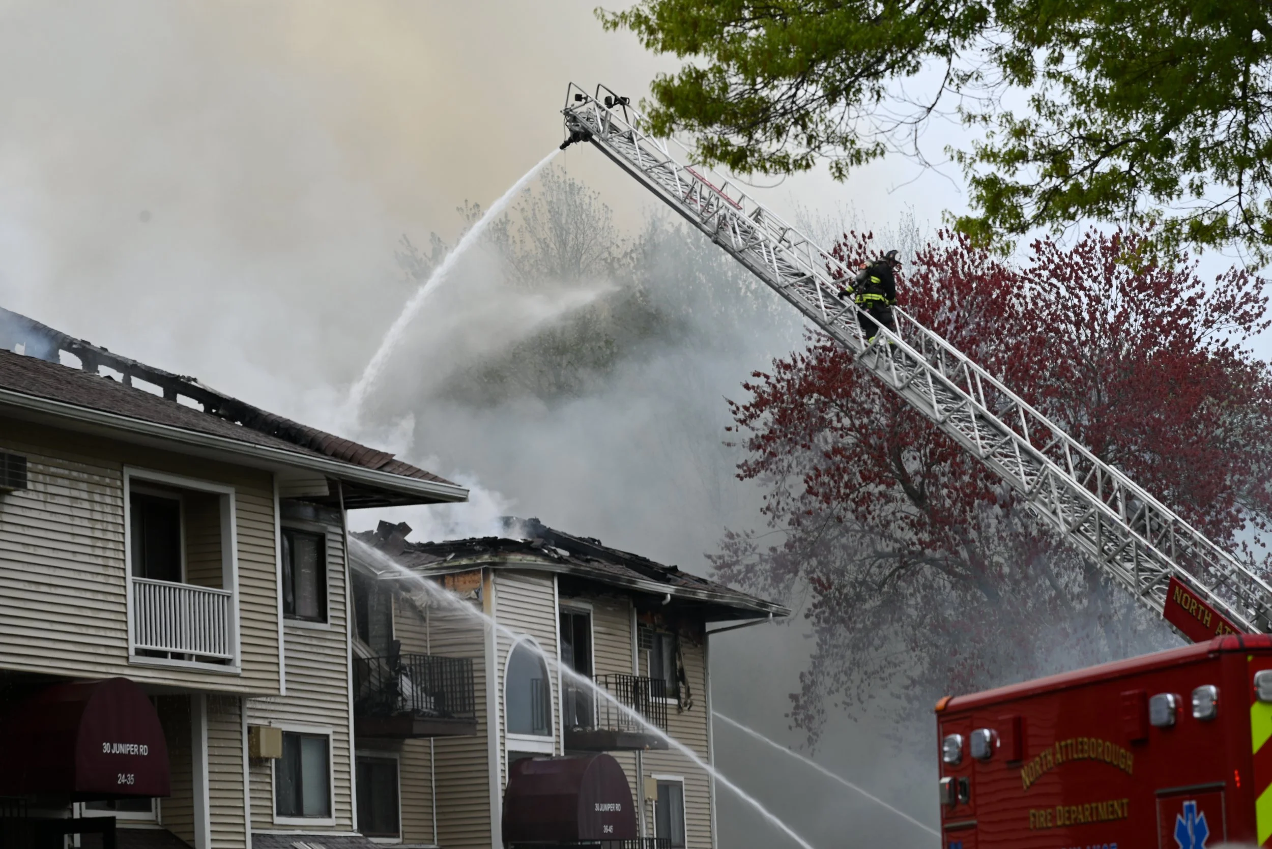 Firefighters use an extended ladder to spray water onto a house fire, with smoke billowing from the roof and flames visible inside.