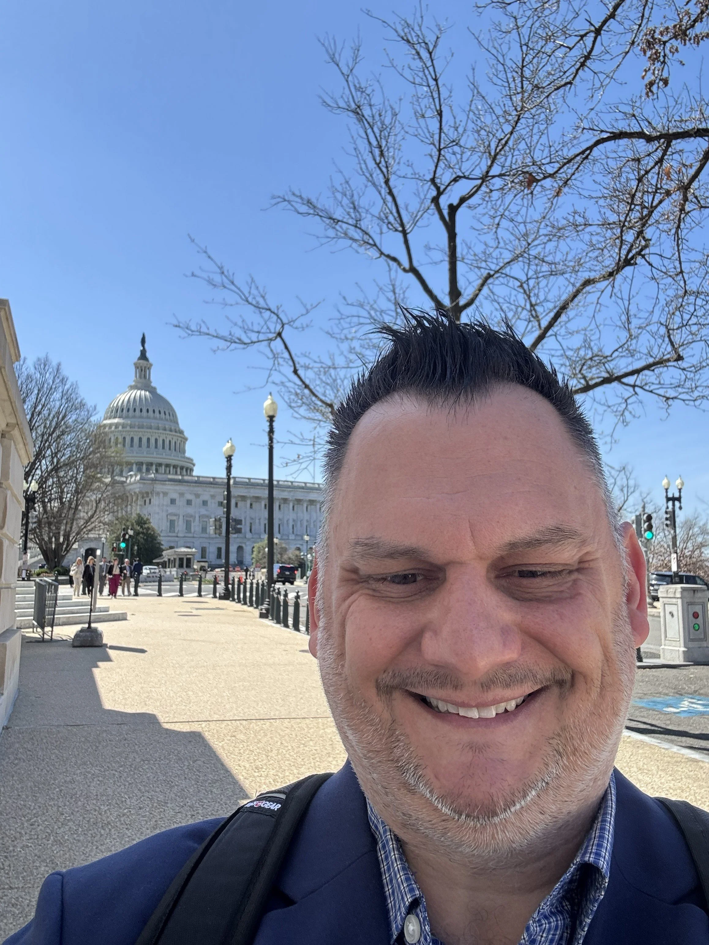 A man smiling in front of the United States Capitol building with a clear blue sky.