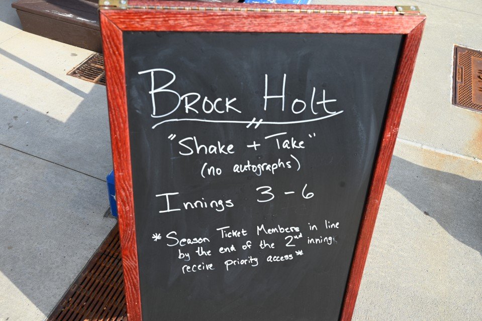 A black chalkboard sign framed in wood advertising Brock Holt, with the slogan 'Shake + Take' (no autographs), showing a range of 3 to 6 Innings, and a note about season ticket members receiving priority access at the end of the second inning, placed outside on a sidewalk.