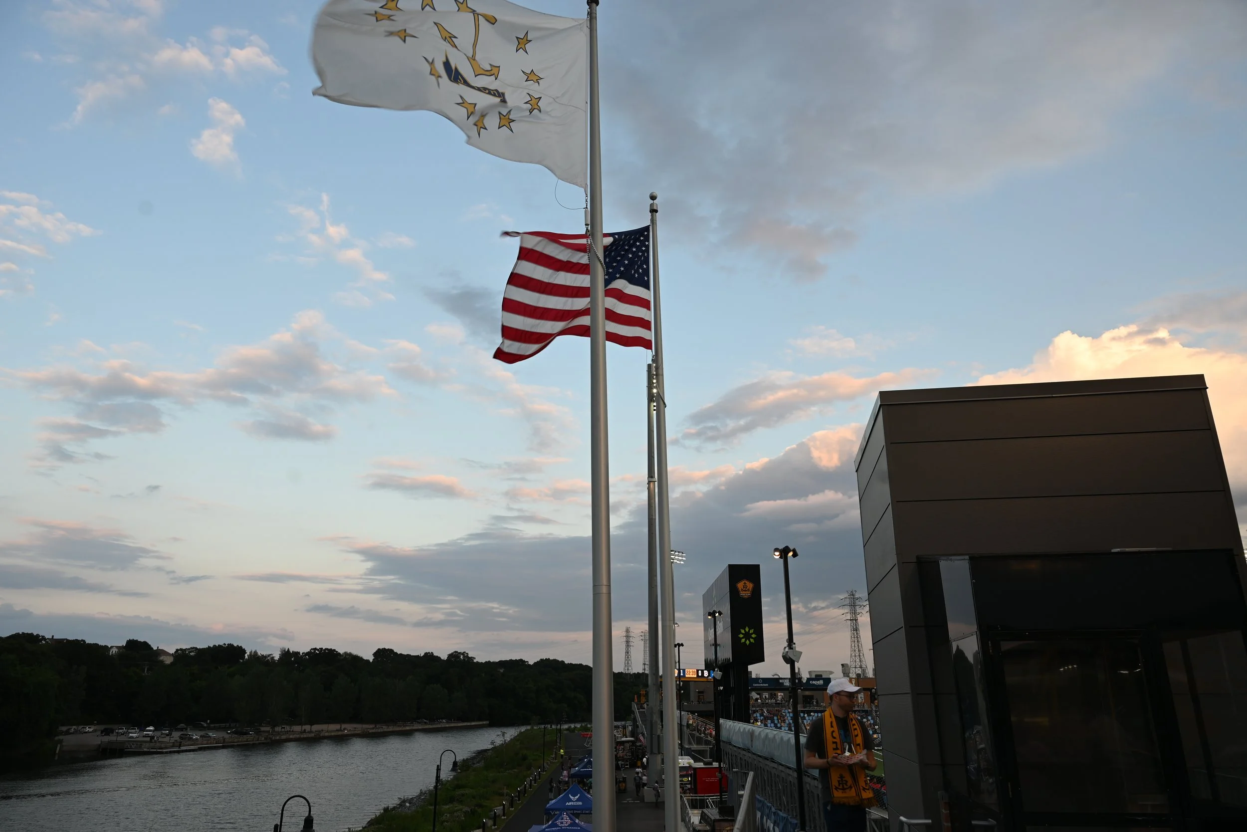 Three flags flying on flagpoles along a riverwalk at sunset, with a man wearing a yellow and black scarf near a modern building and a sports stadium in the background.