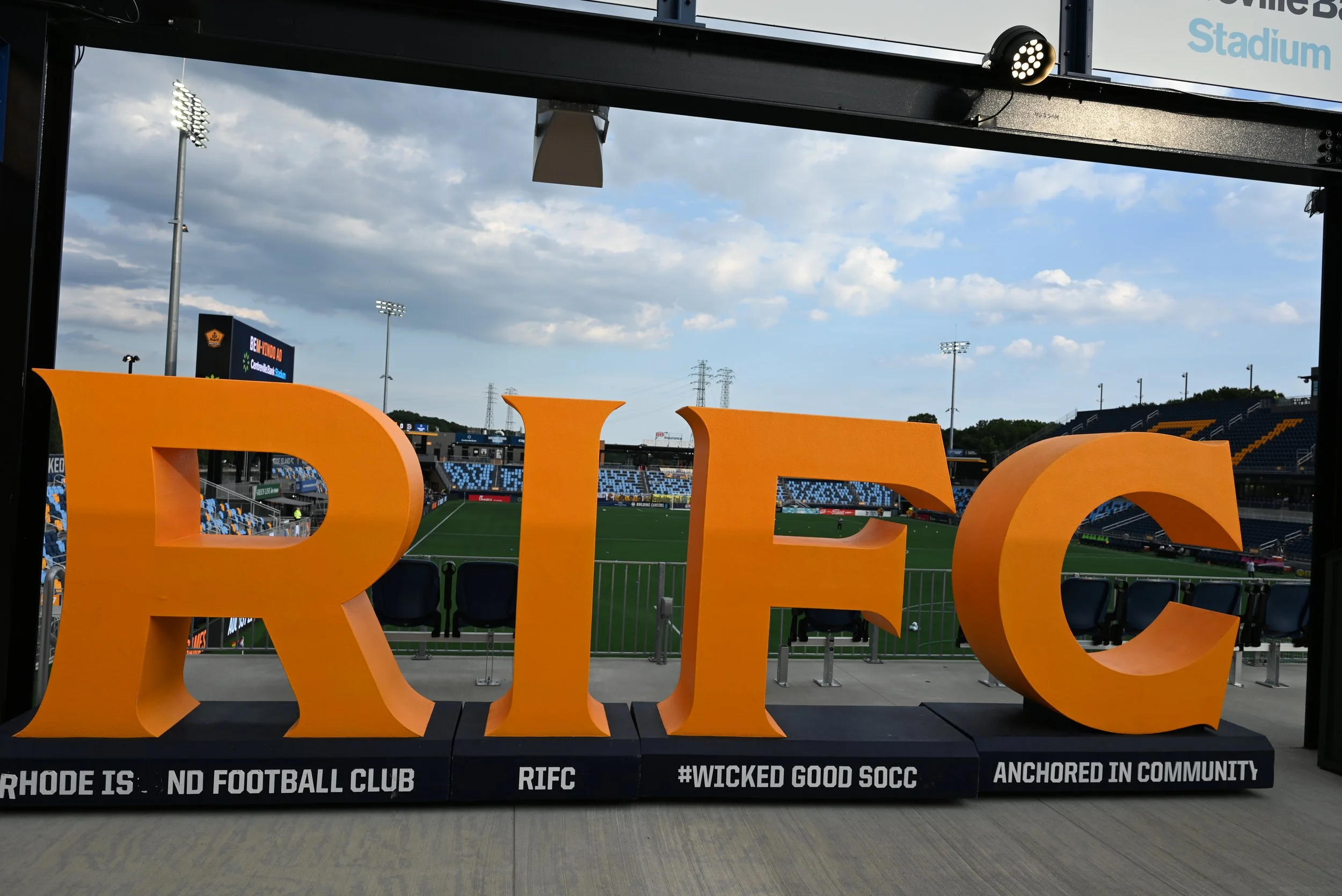 Large orange letters forming the word 'RICH' on a stadium viewing platform with a football field, stands, and floodlights in the background under a partly cloudy sky.