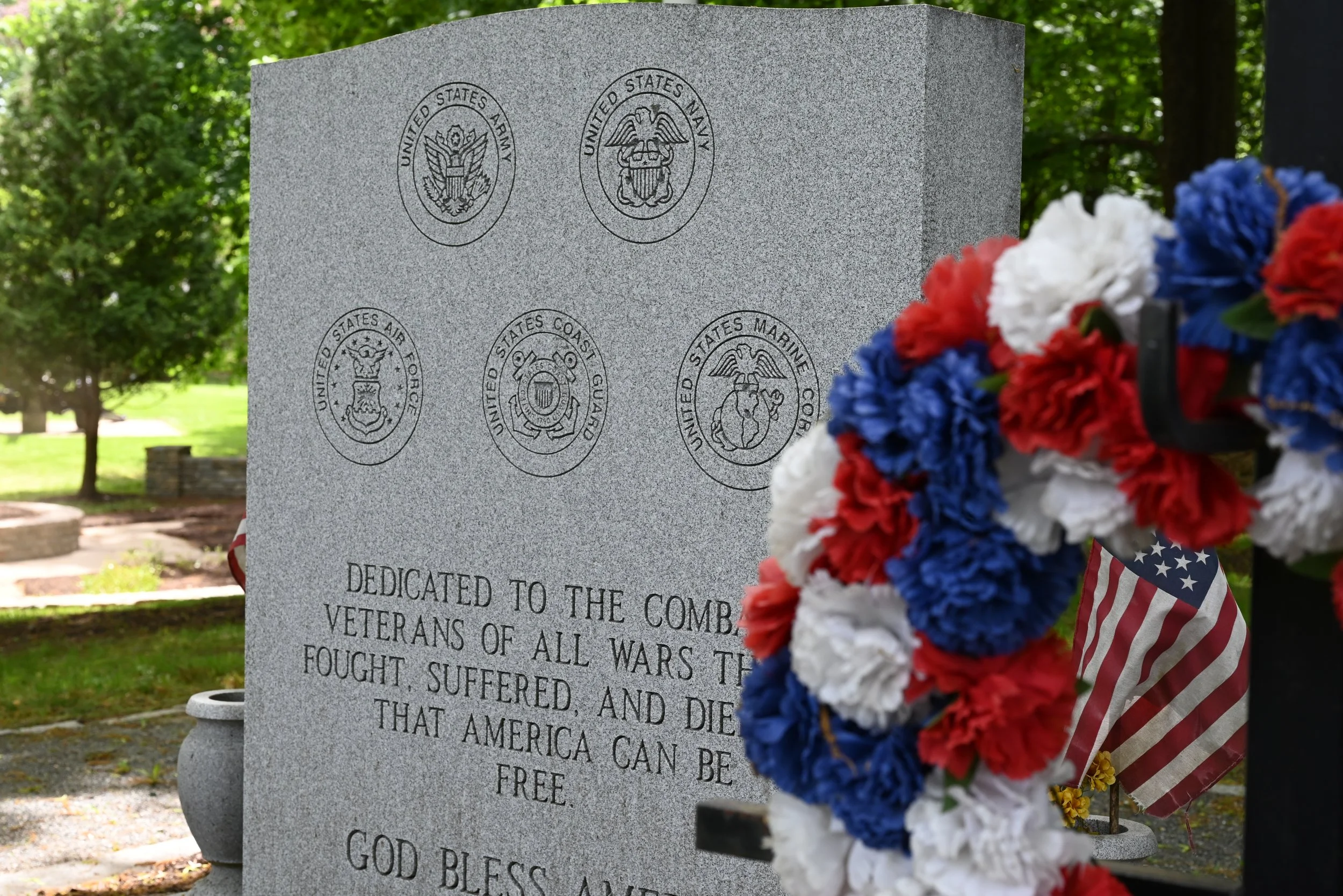A gray granite memorial with engraved seals of five U.S. military branches and a wreath of red, white, and blue flowers in the foreground.