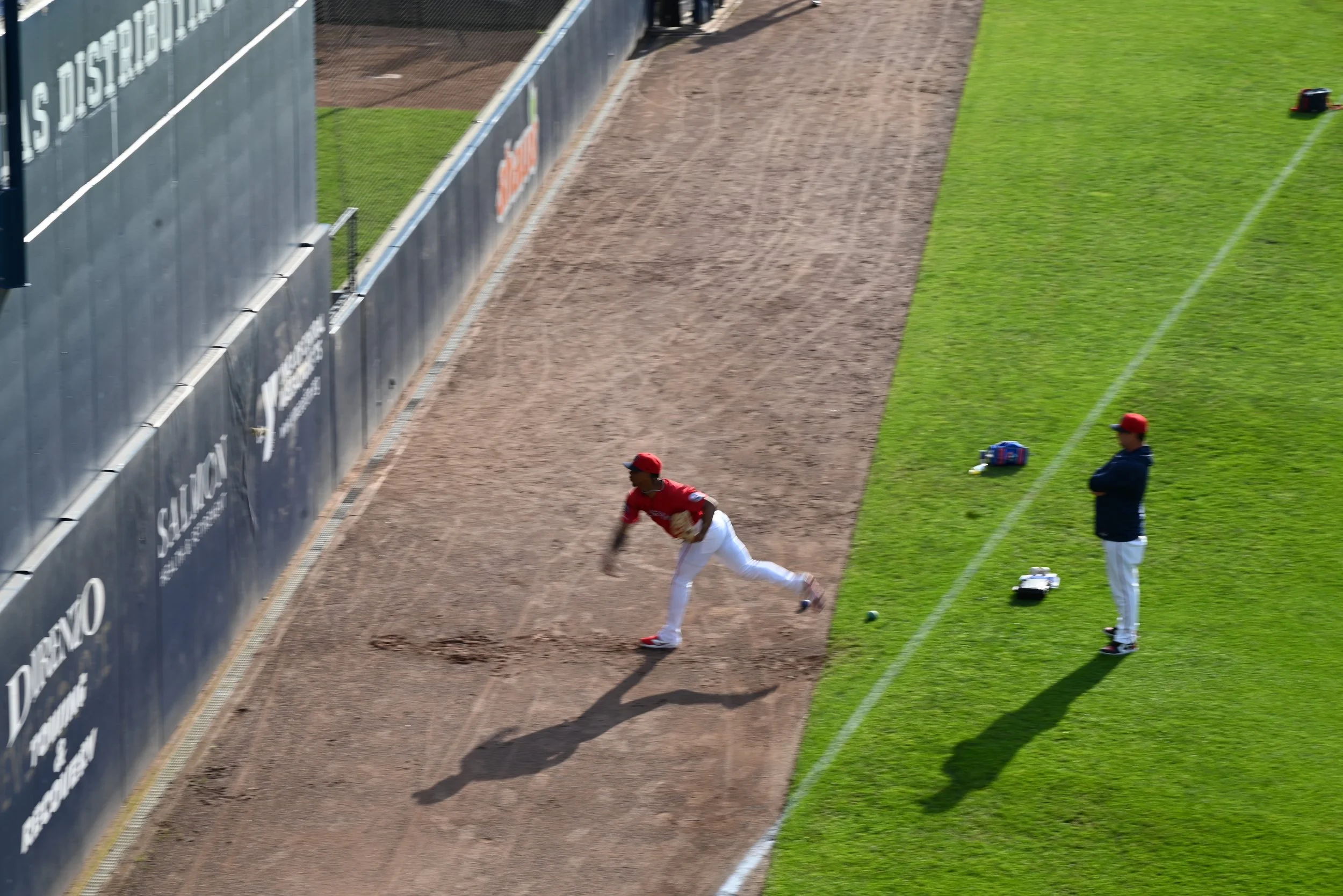 A baseball player in a red jersey and white pants slipping on the ground near the outfield fence, with a person in a dark jacket and red cap standing nearby on the grass.