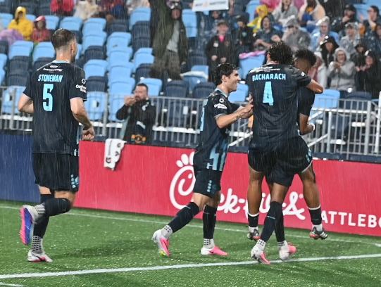 Soccer players celebrating on the field during a match, with fans in the stands and rain falling.