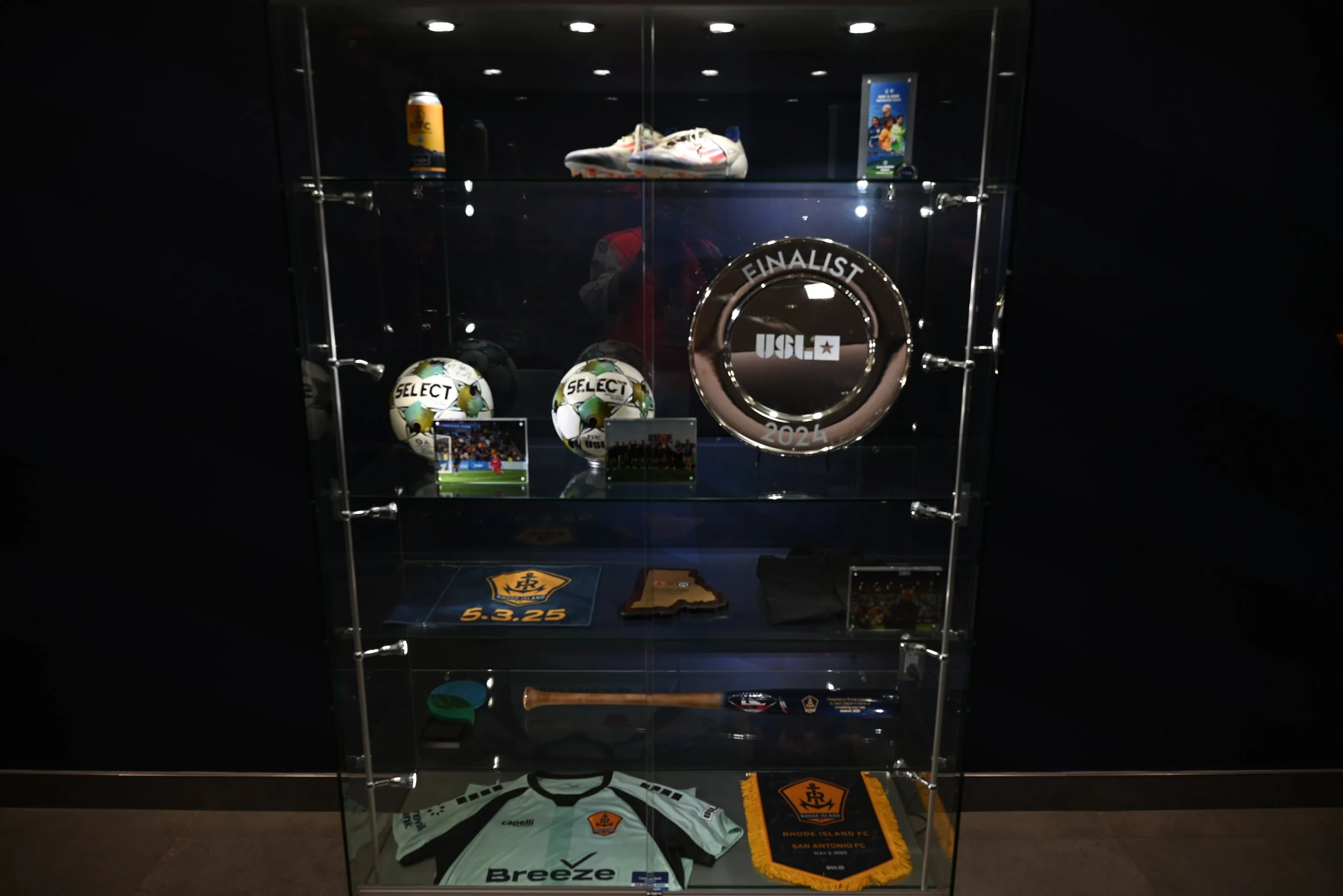 Display case with soccer memorabilia, including a pair of white and red Nike soccer shoes, matching soccer balls with a yellow and green design, and various team and event items associated with USL FINALIST 2024, Rhode Island FC, and San Antonio FC.