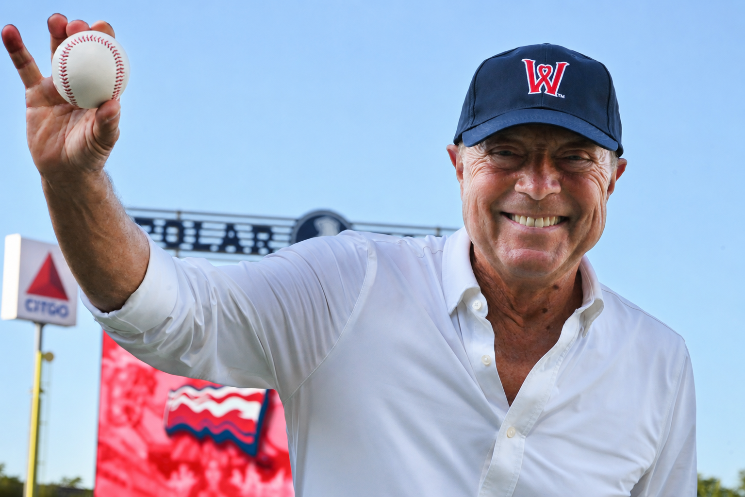 An older man smiling while holding a baseball in his right hand, outdoors on a sunny day with signs and a blue sky in the background.