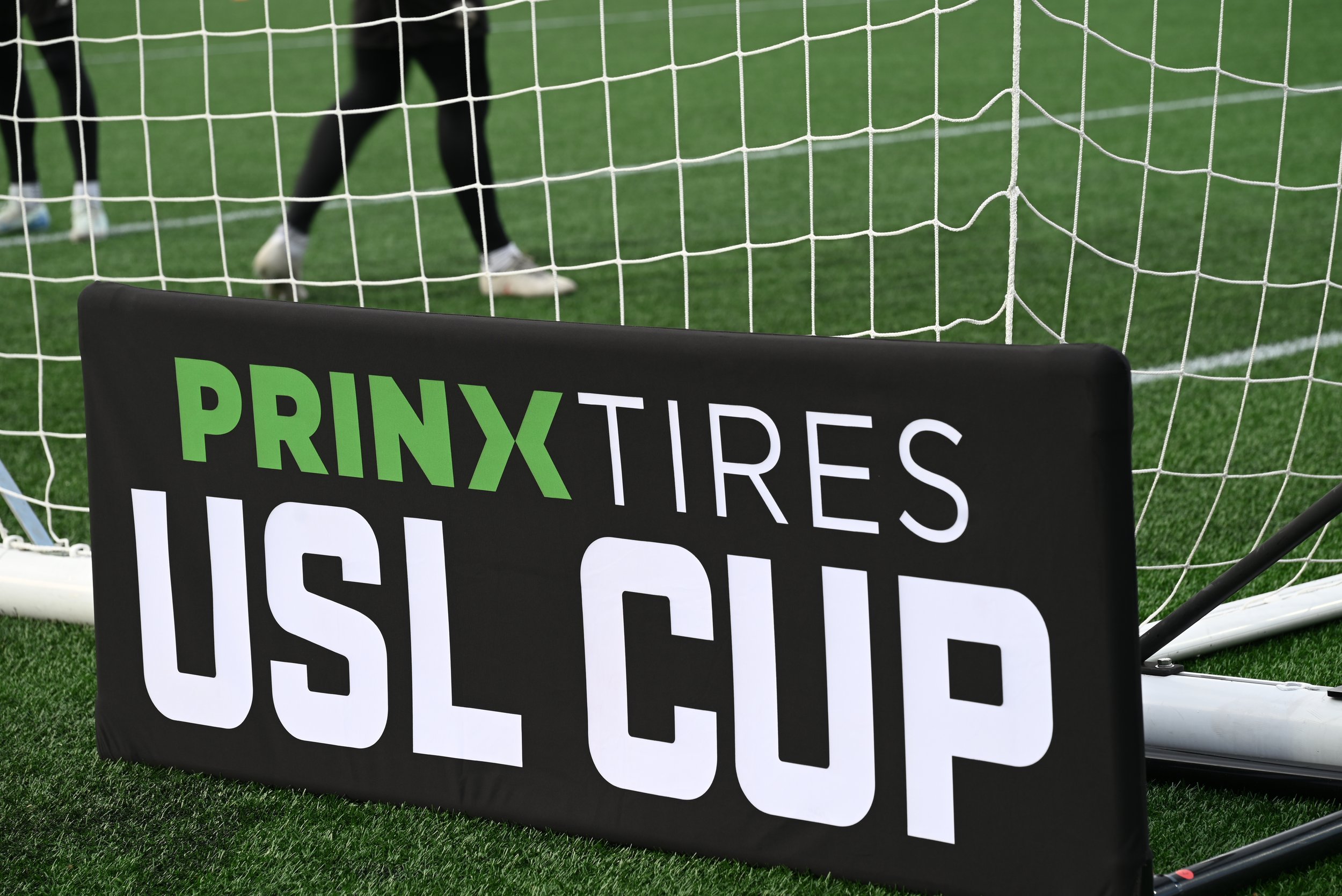 A soccer goal net with a protest sign in front of it that reads "PRINX TIRES, NO OIL CHANGE." In the background, people are visible on a green field.