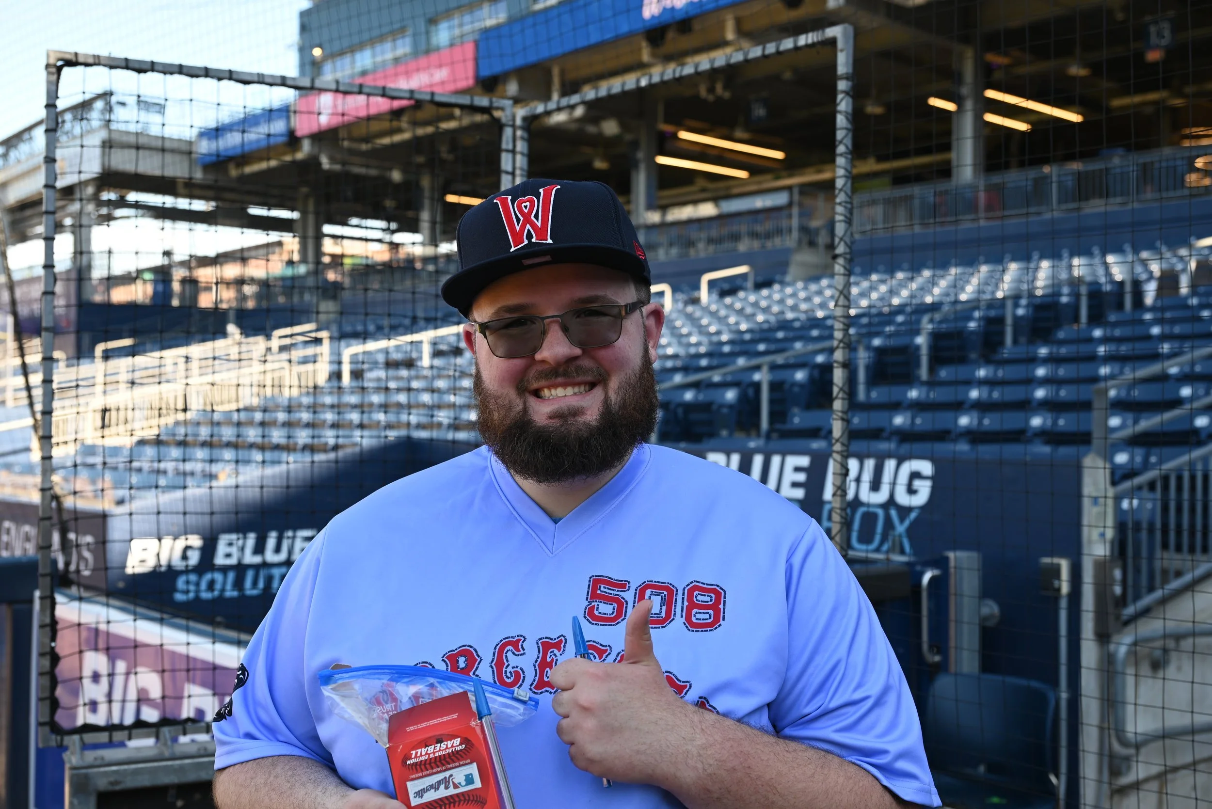 A man with a beard and glasses wearing a baseball cap and a white shirt giving a thumbs up in front of a baseball field fence, holding a plastic bag and a box ofBaseball solution, with stadium seating and a sign for the Big Blue Solution in the background.
