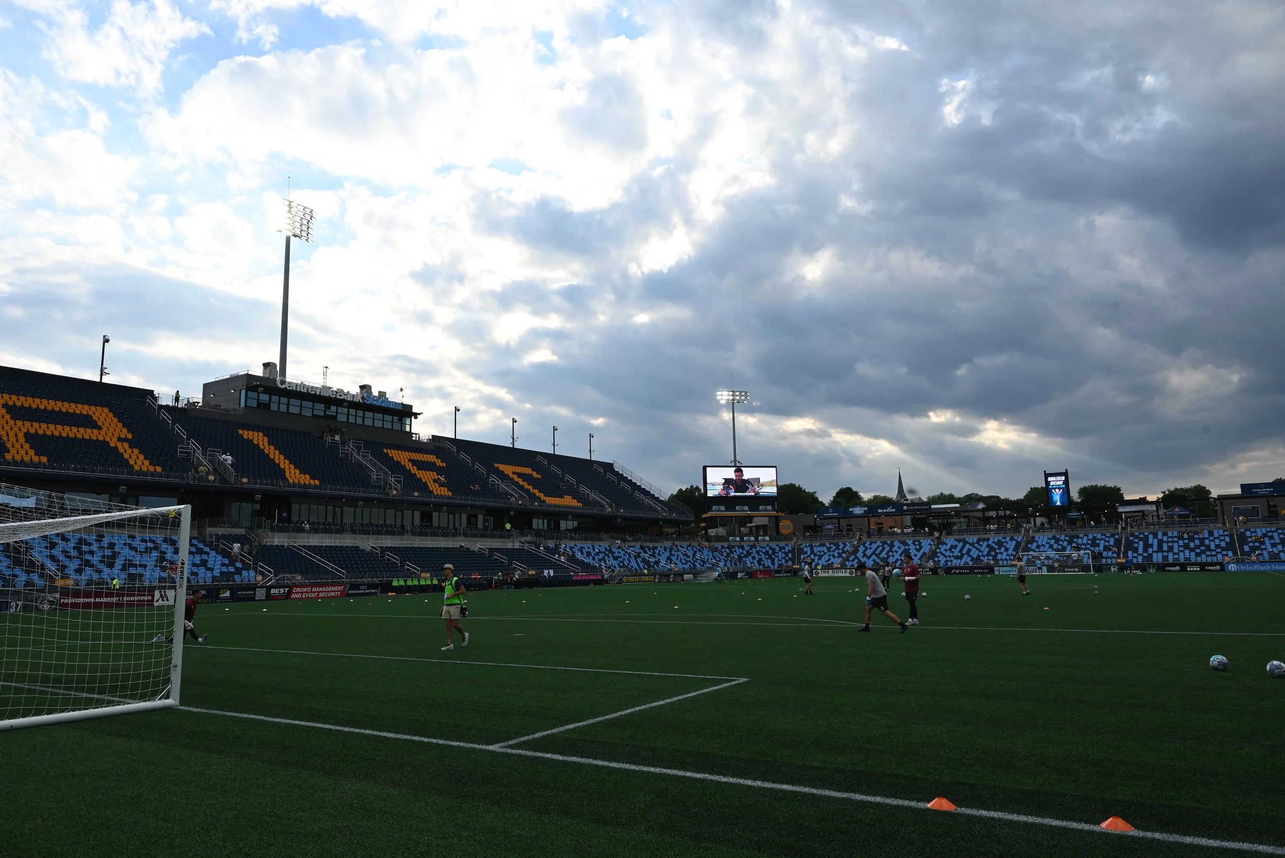 A soccer stadium with players warming up on the field, large screens showing game footage, and mostly cloudy sky overhead.